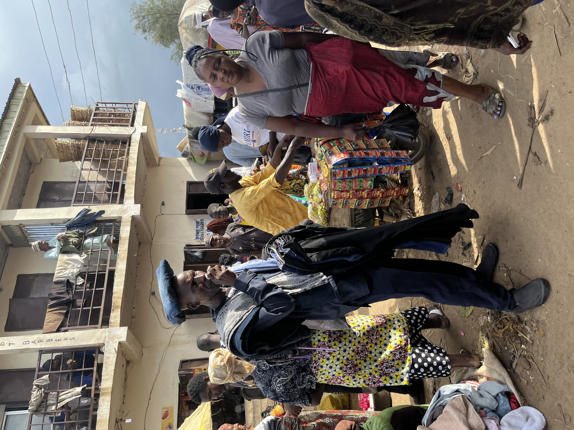 This image captures a bustling scene in Rafin Par, Nigeria, likely a market given the array of goods and people. Several individuals are visible, engaged in various activities. In the foreground, a man in a dark jacket and blue head covering appears to be speaking or gesturing, holding a dark object to his mouth. Beside him, a woman in a colorful, patterned skirt and a yellow and black patterned dress is also present. Further back, a woman in a grey t-shirt and red skirt is interacting with a vendor, whose stall is laden with small packaged goods. Another man in a yellow shirt stands near the stall. The background shows buildings with balconies and windows, some with bars. Baskets of what appears to be produce are stacked on a balcony. The ground is dusty and strewn with debris. The lighting suggests it is daytime, with bright sunlight casting shadows. The overall atmosphere seems lively and active, indicative of commerce and community interaction. There is some visible text on a sign, partially obscured, that reads "MONIE POINT 5156182888".