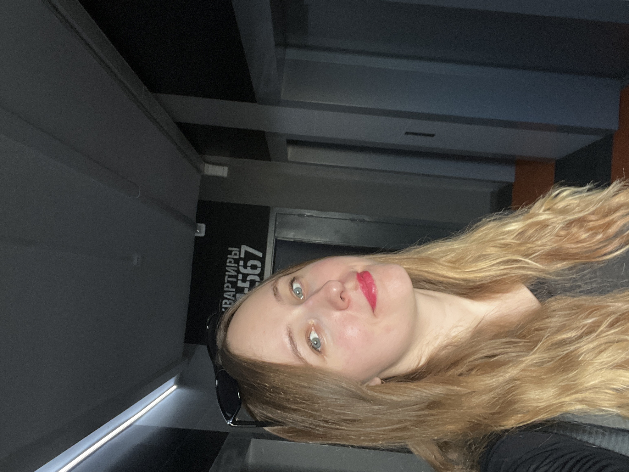The image shows a woman with long, wavy blonde hair and blue eyes, wearing red lipstick and black sunglasses. She appears to be indoors in a modern, commercial or residential building. In the background, there is a sign in Russian: "квартиры 567" (apartments 567). The architectural features are contemporary, with sleek lines and a neutral color palette. Based on the Russian writing, the location is likely in Russia. It is not possible to definitively determine the specific location in Mytishchi, Russia, without more context.