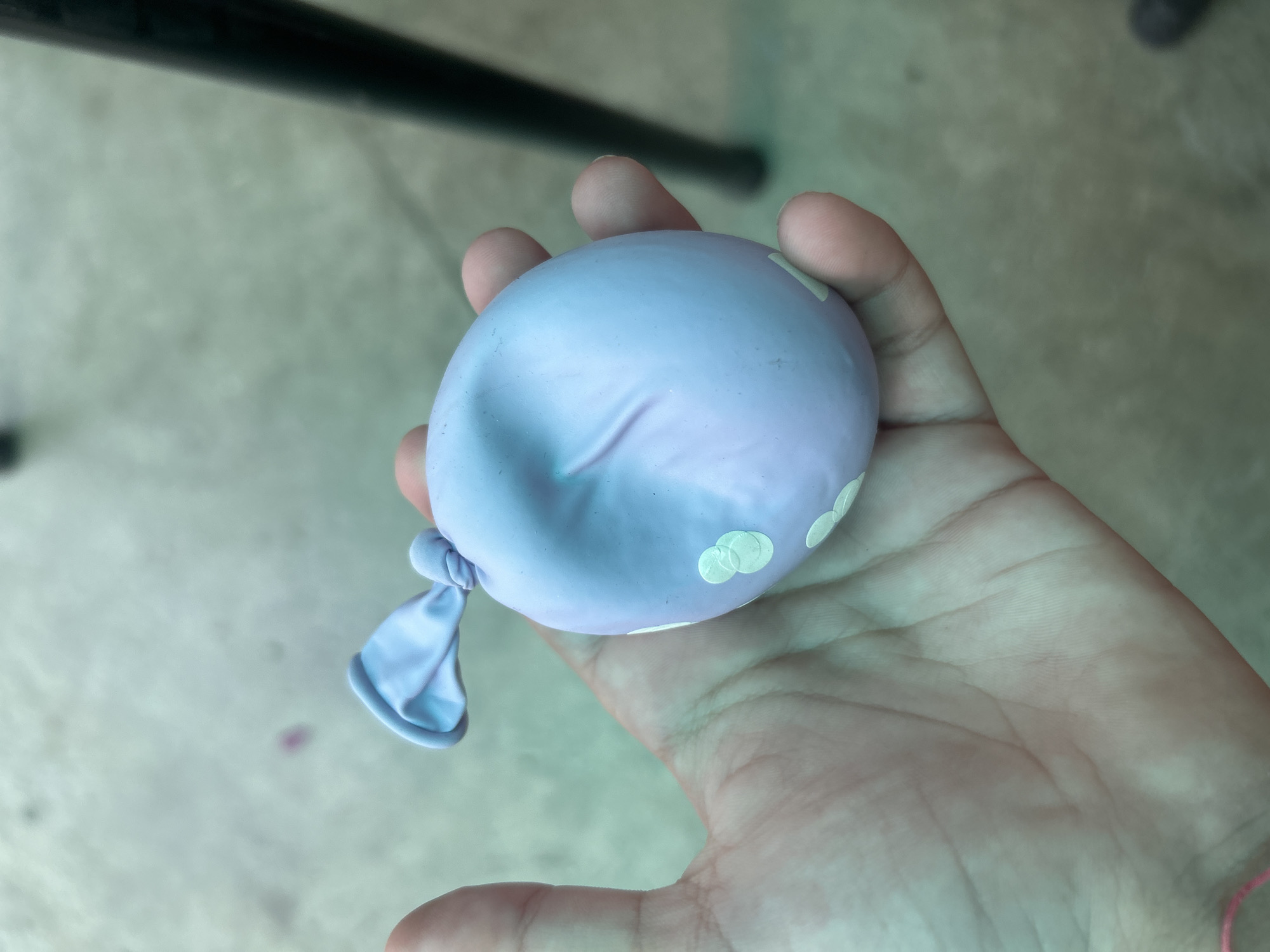 A person's hand is holding a deflated, light purple water balloon with white circular patterns. The balloon is knotted at the top. The background appears to be an outdoor surface, possibly concrete or pavement, with some blurry elements that suggest a natural or man-made environment. There is no discernible location in the city of Govedare, Bulgaria.
