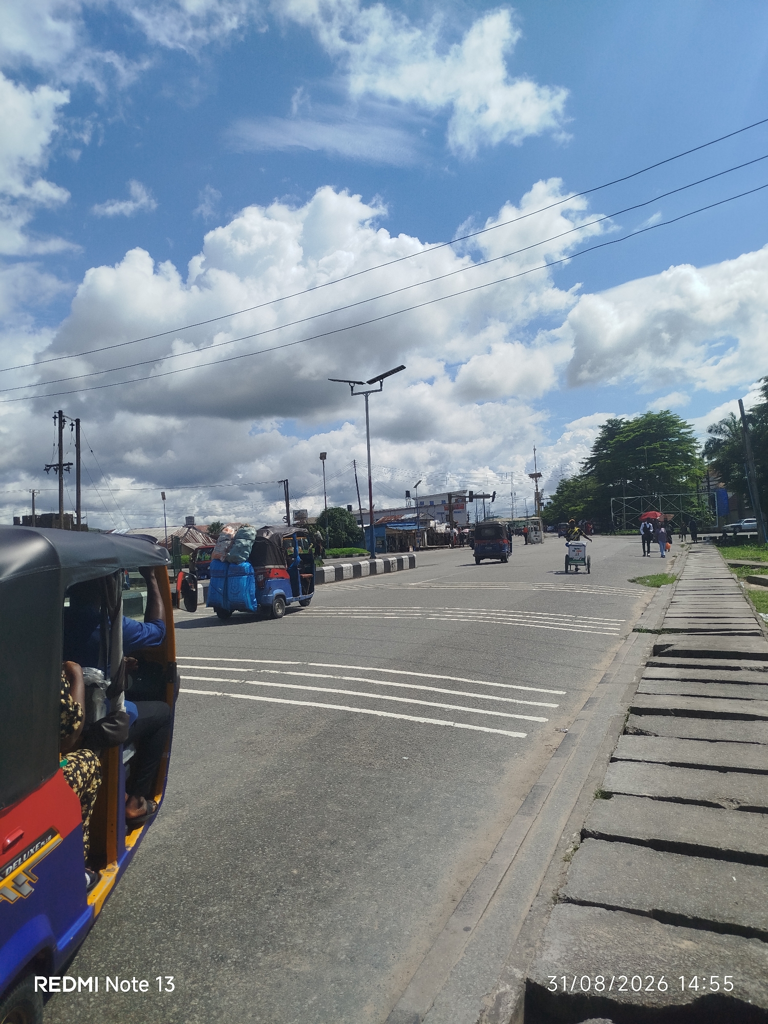 In Yenegoa, Nigeria, this image captures a street scene on a cloudy day. A blue and red auto-rickshaw, loaded with luggage, is moving down the road. In the foreground, another auto-rickshaw is partially visible, with passengers inside. The road is paved and has white lane markings. To the right, a concrete sidewalk with cracked sections runs alongside the road. In the background, trees and buildings are visible, along with utility poles and streetlights. The sky is a mix of blue and white clouds. The timestamp "31/08/2026 14:55" and "REDMI Note 13" are visible in the lower right and left corners respectively.