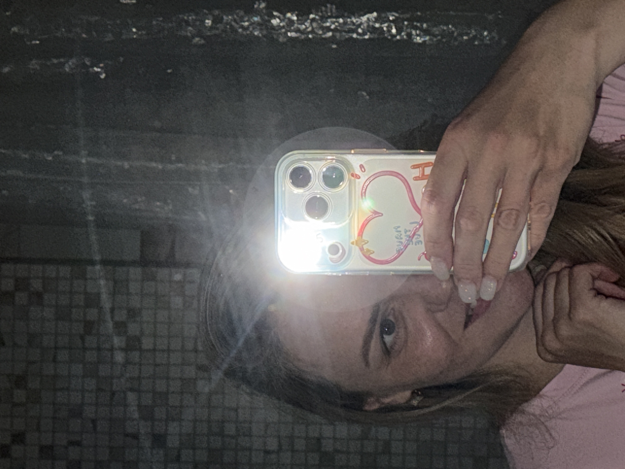 A woman takes a selfie in front of a dark, reflective surface with a tiled pattern. The reflection shows a dimly lit, possibly urban, environment. The woman is holding a smartphone with a decorative case, its flash creating a bright glare. She is partially visible, with light skin and a pink shirt. The background appears to be textured or uneven. There is no verifiable information to determine if the location is in the city of Votkinsk, Russia.