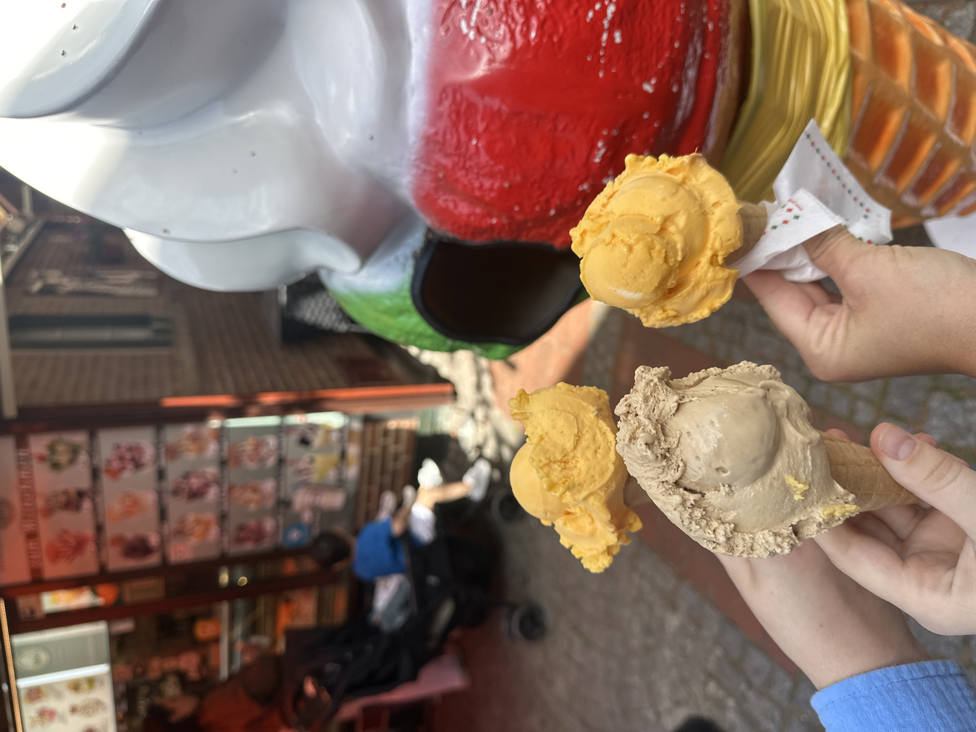 Here is a description of the image:
The image depicts an eye-level, outdoor scene with ice cream cones. Two people are holding ice cream cones aloft. The ice cream cones are of different colors; one is light brown, and the other is a yellow-orange hue. There is a child with a stroller standing in front of a storefront with pictures on the wall. The foreground includes a part of an oversized clown head that is painted with white, red, green and yellow coloring. The photograph was taken at the entrance of the "Eis Cafe Capri", located in Buxtehude, Germany.