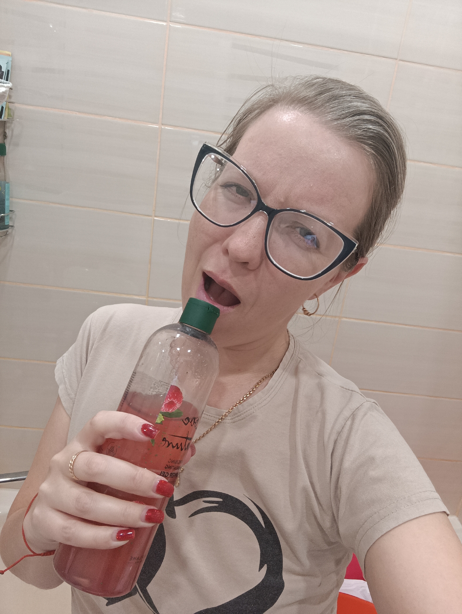 A woman with fair skin, light-colored hair, and black-rimmed cat-eye glasses is captured in a close-up, likely in a bathroom setting due to the light-colored tiled walls. She wears a light beige t-shirt with a dark abstract design, a gold-colored chain necklace, and a gold ring on her left ring finger. Her fingernails are painted red, and a red string bracelet is visible on her left wrist.
The woman is holding a clear plastic bottle filled with a pinkish-red liquid, featuring a dark green cap, to her open mouth. Her expression is animated and playful, with her eyes wide. The bottle's label displays a prominent graphic of a raspberry, and mirrored text on it includes the word "RASPBERRY," indicating the product's flavor or ingredient. Other mirrored words like "МНЕЕ" (less) and "ЧЕРРИ" (cherry) are faintly visible.
The activity appears to be a lighthearted or humorous pose, pretending to drink directly from the product bottle. To the left, a shelf with toiletries and towels is partially visible. The scene is illuminated by indoor lighting. This photograph was taken in Chaikovsky, Russia.