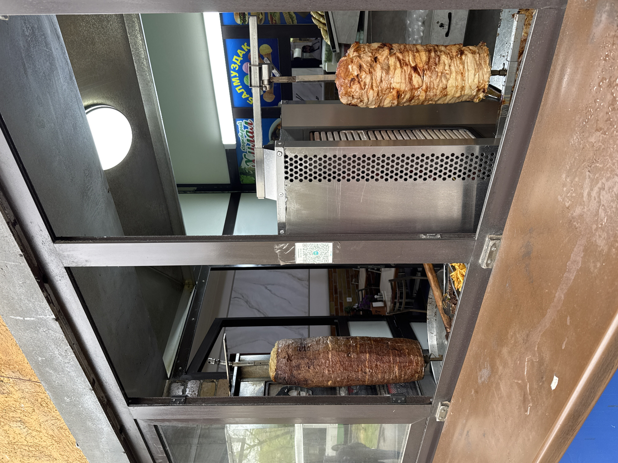 The image captures the interior of a food stall or kiosk, likely viewed through a service window or opening, in Bishkek City, Kyrgyzstan. Two vertical rotisseries dominate the scene, each holding a large cone of meat actively cooking for döner kebab or shawarma. The upper rotisserie cooks lighter-colored meat, likely chicken or turkey, while the lower one prepares darker meat, possibly beef or lamb.
The equipment features stainless steel construction with perforated panels. A bright, circular white light fixture is visible on the ceiling. In the background, a blue sign with white and yellow Cyrillic text is prominently displayed. Visible text includes "БАЛМУЗДАК" (Balmuzdak, meaning "ice cream" in Kyrgyz), and fragments like "КАНАТУУ" (Kanatuu, possibly related to poultry or "winged") and "АЯЛ" (Ayal). A small, indecipherable QR code sticker is affixed to a glass pane near the top center.
The stall's structure consists of dark metal framing and glass panels. A textured, light-colored exterior wall is seen on the far left, and a brown, textured surface, possibly a counter or another part of the stall, is on the far right. No people are visible, and the scene depicts a functional commercial food preparation area, likely for takeaway or quick service.