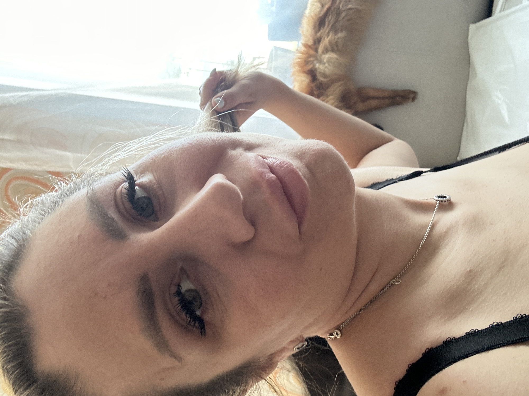 A close-up, selfie-style shot captures a woman with blonde hair pulled back, holding a strand of her hair with her right hand. She is wearing a black bra with lace detailing and a silver necklace. Her makeup includes dark eyeliner and mascara. In the background, an orange cat is visible, lying on a white surface. The scene appears to be indoors, with a window and sheer curtains providing natural light. The woman's expression is neutral. The photograph was taken on April 11, 2023, in Novoaltaysk, Russia.