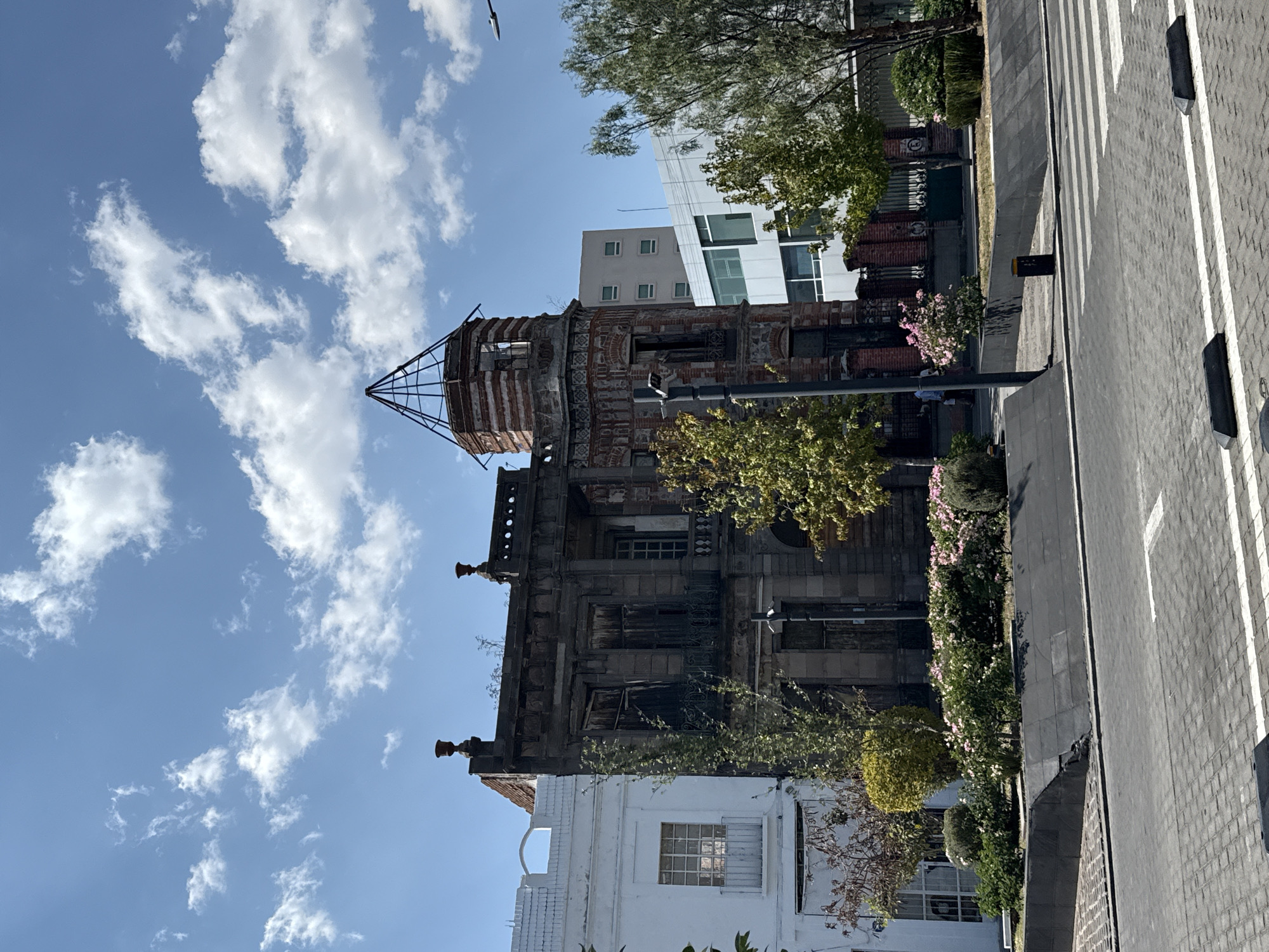 The image shows a tall, weathered building in Puebla, Mexico. The structure has an ornate, decaying facade with a skeletal framework on top, possibly the remains of a dome or tower. The building exhibits signs of age and neglect. In the background are modern buildings. In the foreground, the street has a crosswalk and landscaping. There is no visible human activity in the immediate vicinity.