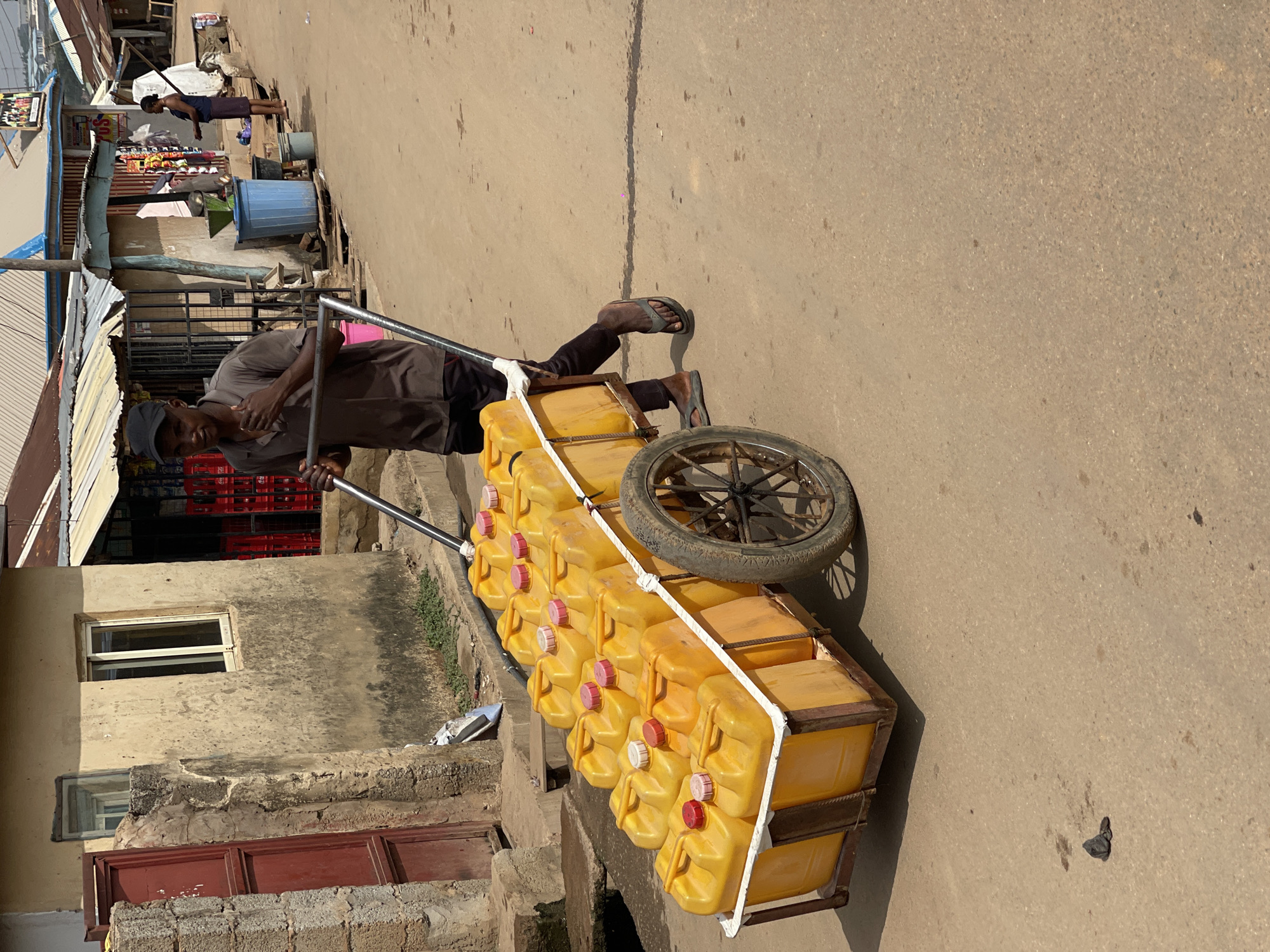 The image depicts a bustling street scene in Kuchingoro, Nigeria, centered on a man pushing a heavily loaded cart. The man, with dark skin, is dressed in a short-sleeved brown shirt, dark trousers, and flip-flops, and appears focused on his task. He is propelling a robust metal cart, which features at least one visible spoked wheel on its right side. This cart is filled with numerous bright yellow plastic jerrycans, each sealed with either a red or white screw cap. These containers are secured firmly to the cart with white ropes or straps, indicating they are likely transporting water or other liquids.
Along the left side of the street, a series of informal market stalls or small shops line the path. These structures are characterized by their corrugated iron roofs and weathered concrete walls. Visible items include what appear to be fruits displayed at one stall and stacks of red crates at another. In the background, a second individual, seemingly a woman, stands near these market areas. A concrete drainage channel runs alongside the street, separating the pathway from the buildings.
The scene is illuminated by bright natural daylight, suggesting it is midday or early afternoon, with distinct shadows cast across the ground and structures. The street surface itself appears unpaved and worn, showing an uneven texture and a noticeable crack or line down its center. The overall impression conveys a sense of daily commerce and labor in an active, semi-urban setting. No specific emotions are discernible from the man's expression. A small, blurry sign in the background, on one of the stalls, might display the text "SOS," but it is indistinct.