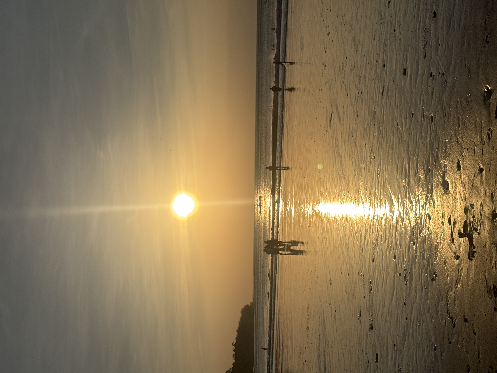 The image depicts a beach scene in Kuta Selatan, Indonesia, during sunset. The sun, a bright, luminous orb, is positioned high in the sky, casting a warm, golden glow over the entire scene. A prominent sunbeam extends vertically downwards from the sun. The sky is a gradient of pale yellow and grey, with wispy clouds. The ocean stretches across the middle ground, with gentle waves lapping at the shore. Several individuals are visible on the beach and in the shallow water. In the foreground, the wet sand of the beach reflects the sunlight, creating a shimmering, elongated reflection of the sun. Small pebbles and indentations are scattered across the sand. In the middle ground, a group of three people stand in the shallow water, their figures silhouetted against the bright light. Further out in the water, other individuals can be seen, some appearing to be surfing or wading. A boat with a canopy is visible in the distance on the ocean. The background features a dark, tree-covered coastline.