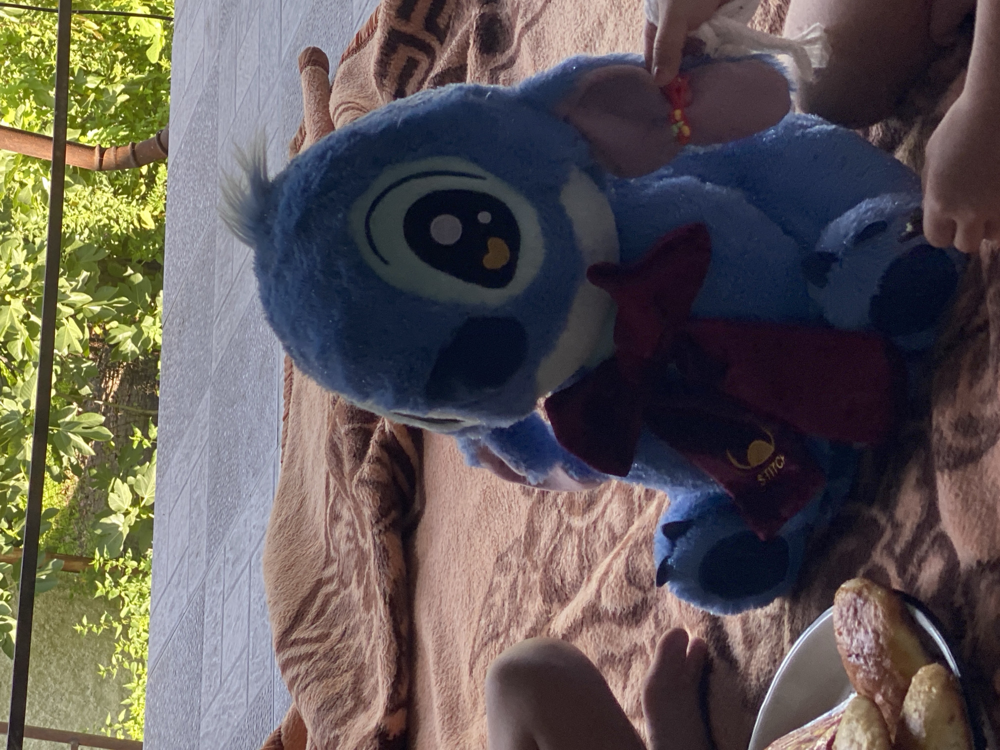 The image depicts a plush toy of Stitch from Disney's Lilo & Stitch, positioned on a brown blanket. The toy is blue with large eyes and is wearing a red bow. To the left of the toy, a person's legs are visible, and to the right, a child's hand is interacting with the toy, holding a bracelet made of colorful beads. A white bowl containing several baked goods, possibly pastries, is also present in the lower left corner. The background shows a tiled surface and green foliage, suggesting an outdoor setting. The scene is likely located in Lagodekhi, Georgia, given the provided location context.