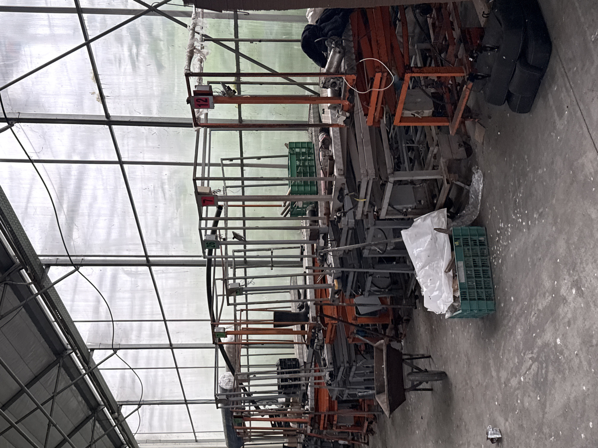 The image appears to depict the interior of a greenhouse or similar structure, possibly in Stara Zagora, Bulgaria, though no specific features confirm the location. The scene is dominated by a large collection of metal scaffolding, carts, and various pieces of equipment, creating a cluttered environment. A green crate and a white bag are present on the concrete floor. The structure features a metal frame and a translucent roof. No people are visible.