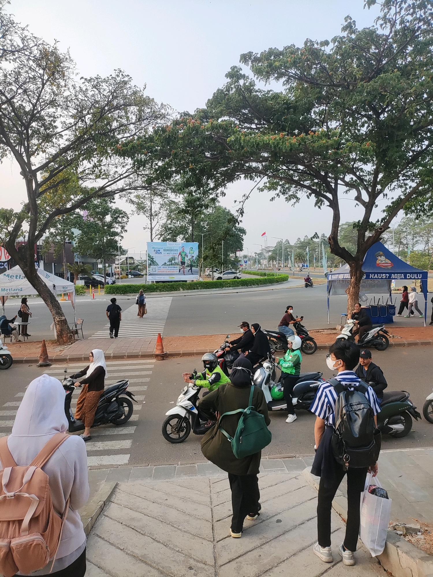 The image depicts a street scene in Telagamurni, Indonesia, likely during daylight hours. A busy intersection with a pedestrian crosswalk is visible, with several people and motorcycles present. In the foreground, a person wearing a white hoodie and a light pink backpack stands on a sidewalk, facing away from the camera. To their left, a woman in a white hijab and brown clothing rides a black motorcycle across the crosswalk. Another person on a white motorcycle, wearing a green jacket and helmet, is also crossing the street.
Further back, a group of people are visible near a blue tent with "HAUS? AQUA DULU" written on it, suggesting a commercial stall. A large banner displaying "Metland" and advertisements for a running event and apartments is prominently displayed across the street. Several other individuals are seen walking or riding motorcycles in the background, navigating the intersection. Trees line the street, providing shade. The overall environment suggests a mixed-use area with commercial activity and residential proximity.