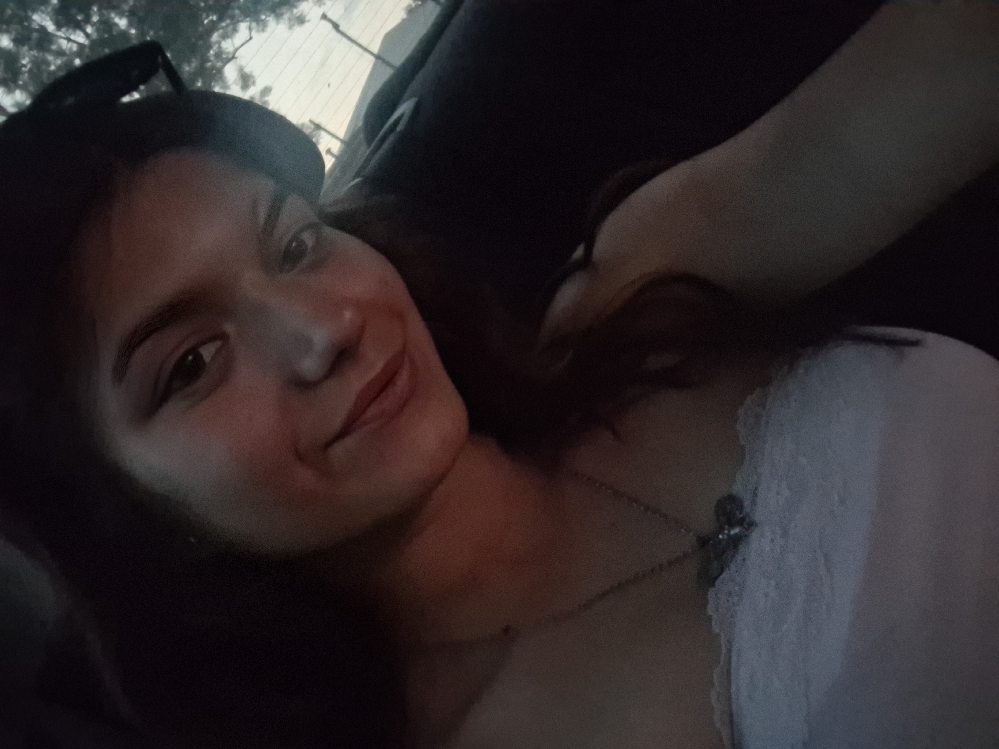 This media file is a close-up, selfie-style photograph of a young woman. She is lying down, possibly in a car given the background which appears to be a window with trees and a sky visible through it, suggesting an outdoor setting. The lighting suggests it is either dawn or dusk, as the light is soft and warm. The woman has dark hair and is looking towards the camera with a slight smile, conveying a relaxed and content emotion. She is wearing a white lace top and a necklace with a pendant. The overall mood of the image is intimate and casual. There is no visible text in the image.