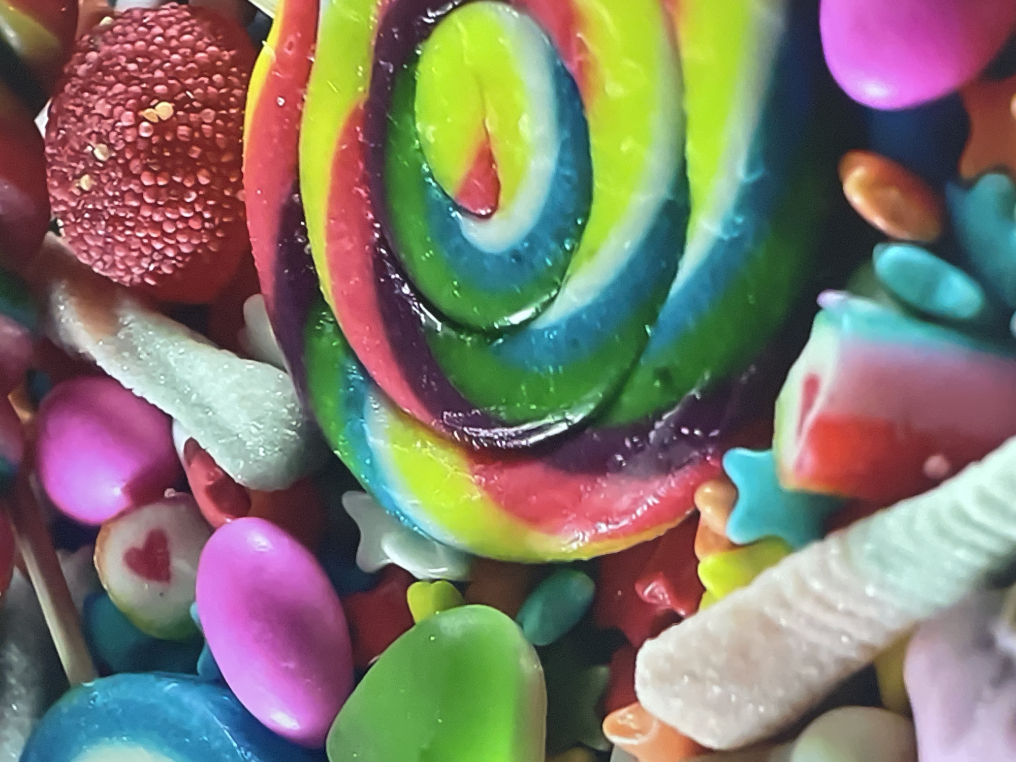 The image shows a close-up view of a collection of assorted candies. A large, colorful lollipop with a spiral design dominates the center. Other candies include small, round candies covered in tiny granules, oval-shaped candies in pink and green, white gummy-like sticks, and star-shaped candies. There is no indication of the location being Rozsoshentsi, Ukraine, or any people or actions in the image. The environment consists solely of the candies themselves.