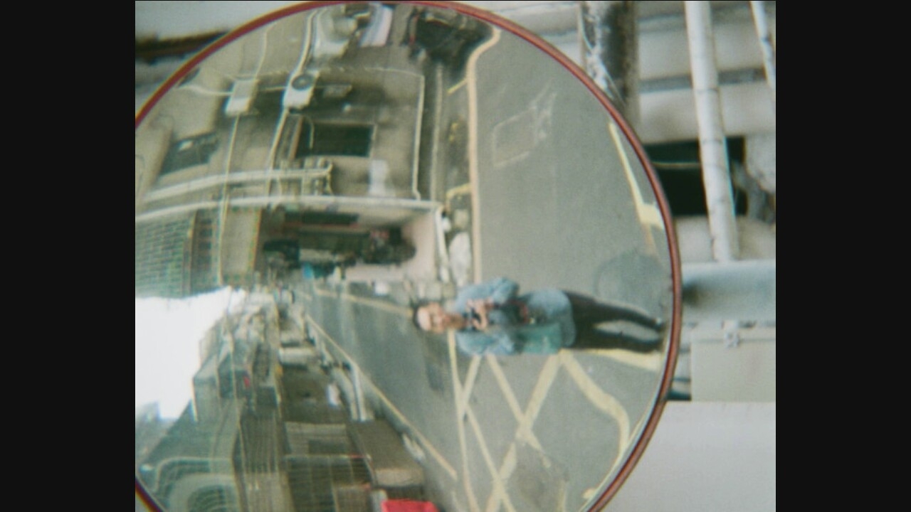 A man stands in a street reflected in a convex mirror, taking a selfie. He is wearing a blue button-down shirt, dark pants, and a crossbody bag. The street appears to be in a city, with buildings lining both sides and a yellow crosswalk painted on the asphalt. The reflection is distorted by the curvature of the mirror, giving a wide-angle view of the surroundings. The buildings are multi-story structures with balconies and windows, typical of an urban environment. The lighting suggests it is daytime.