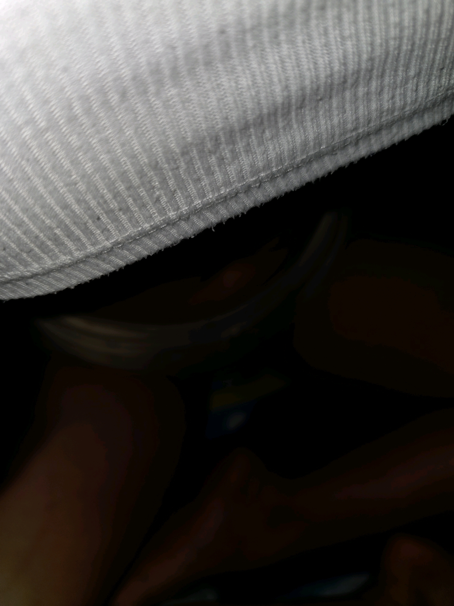 The image shows a close-up view of a person's legs and feet, partially obscured by dark fabric and a white textured surface, possibly a piece of clothing or bedding. The lighting is dim, with some areas appearing illuminated while others are in deep shadow, creating a sense of depth and focus on the skin tones of the legs. The white surface has a ribbed texture, and the dark fabric appears to be soft and draping. The scene is intimate and ambiguous, with no specific location in Phan Thiet, Vietnam, being identifiable.