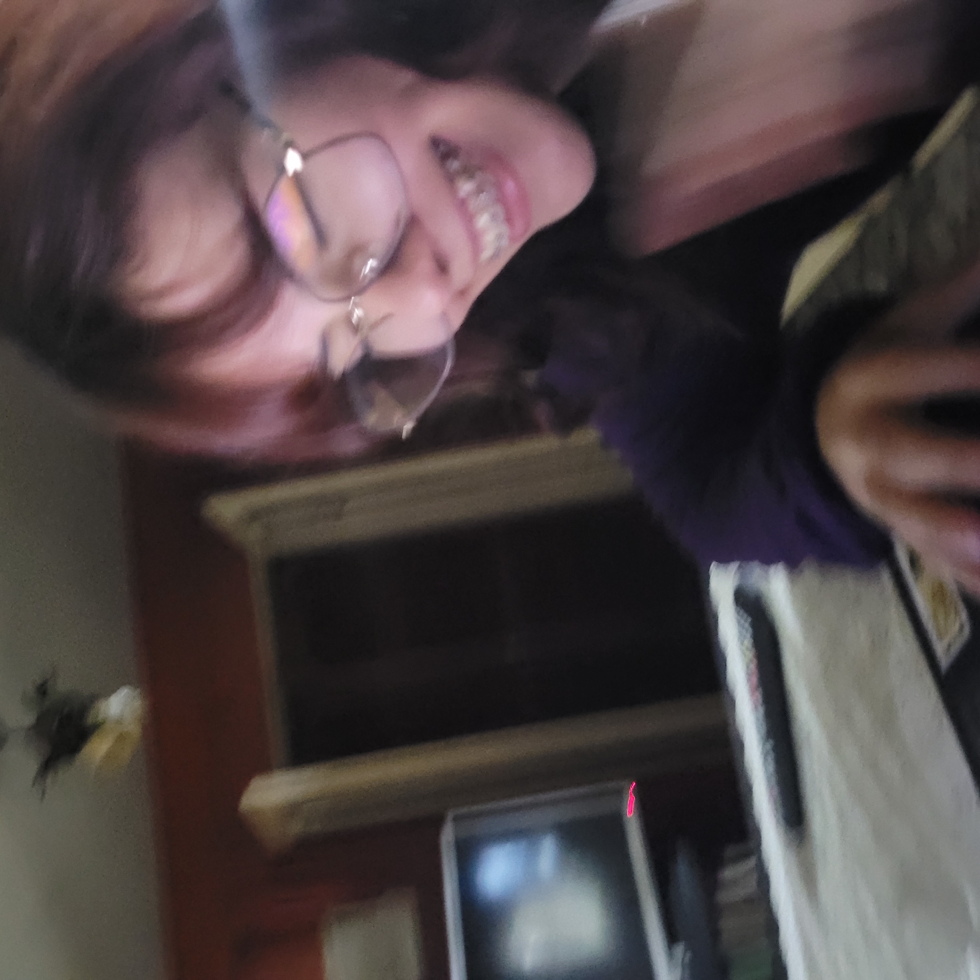 A photograph shows a person with brown hair and glasses smiling with braces, while wearing a purple long-sleeved shirt. In the background is a window, a television, a remote control, and a light-colored object. The photograph may have been taken indoors. The location of the photograph in the city of San Fernando del Valle de Catamarca, Argentina, is not determined.