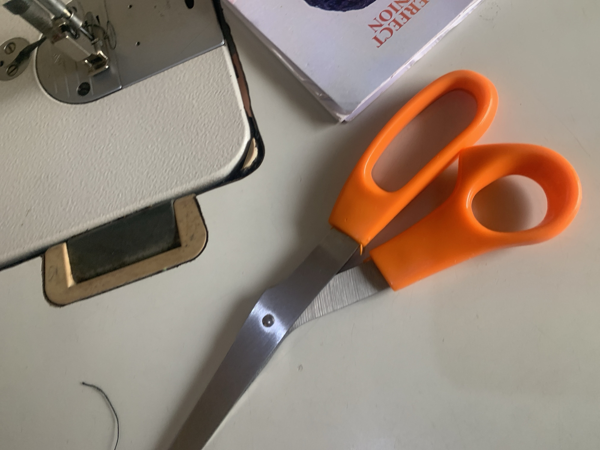 The image shows a close-up, overhead view of a white sewing machine and a pair of orange-handled scissors on a white surface. The sewing machine is partially visible on the right side of the frame, with its needle and presser foot in focus. The scissors are open and positioned diagonally across the left side of the frame, with their blades extended. A thin black thread is visible to the left of the scissors, and a book with text that appears to be "PERFECT UNION" is partially visible beneath the sewing machine. The lighting is bright and appears to be from above. The scene is static and does not depict any action or interaction between subjects. The location is described as Jauro Yinu, Nigeria, but there are no specific visual cues in the image to confirm this location.