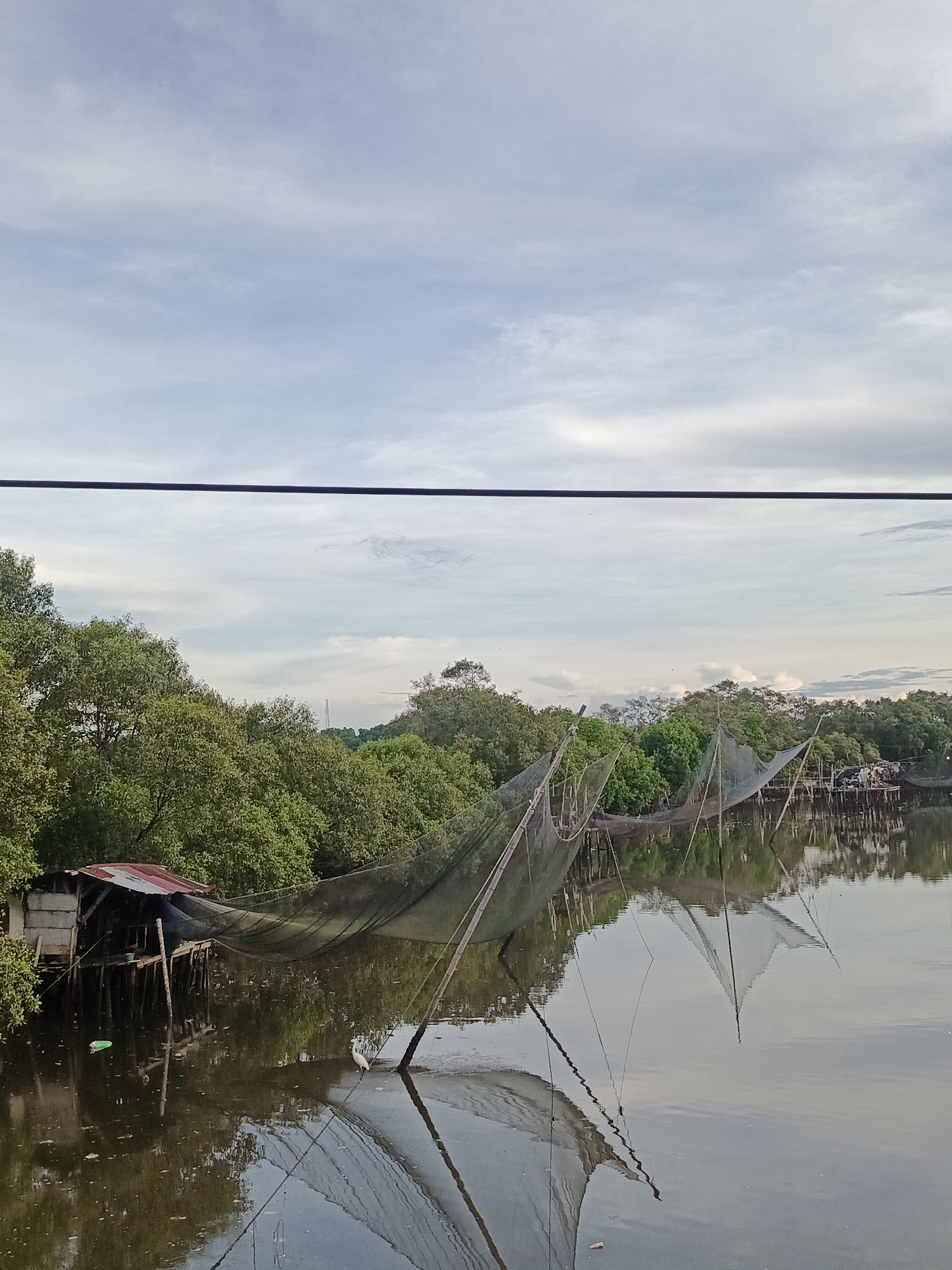 The image depicts a serene waterscape with fishing nets and a small shack in Lhokseumawe, Indonesia. In the foreground, the calm water reflects the sky and the surrounding vegetation. Large fishing nets, supported by wooden poles, are spread across the water, indicating active fishing operations. To the left, a rustic shack with a corrugated metal roof stands on stilts over the water. Lush green trees line the background, creating a natural barrier between the water and the sky. The sky is partly cloudy, with soft white clouds drifting across a pale blue expanse. A thin, dark horizontal line, likely a wire or cable, runs across the upper portion of the image. The scene evokes a sense of tranquility and the traditional way of life in a coastal community.