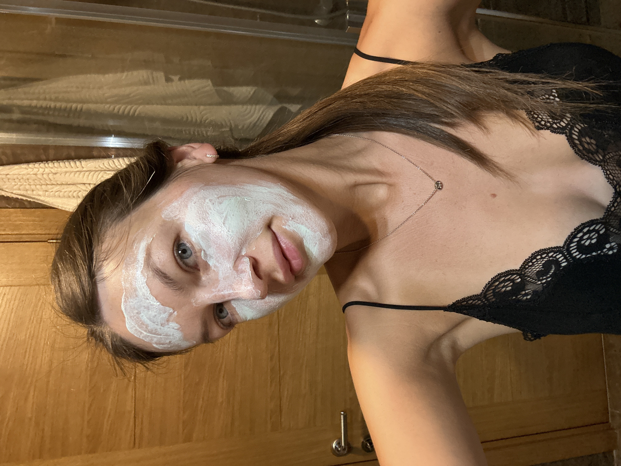 A young woman with blue eyes and a light complexion is shown in a selfie. She has a light green face mask applied to her forehead, cheeks, and nose. Her hair is dark brown and pulled back into a ponytail, with some strands loose around her face. She is wearing a black lace camisole with thin straps. A delicate silver necklace with a small circular pendant is visible on her chest. The background consists of a wooden door on the left and a shower with towels on the right. The scene appears to be in a bathroom. The location is specified as Donji Pobori, Montenegro.