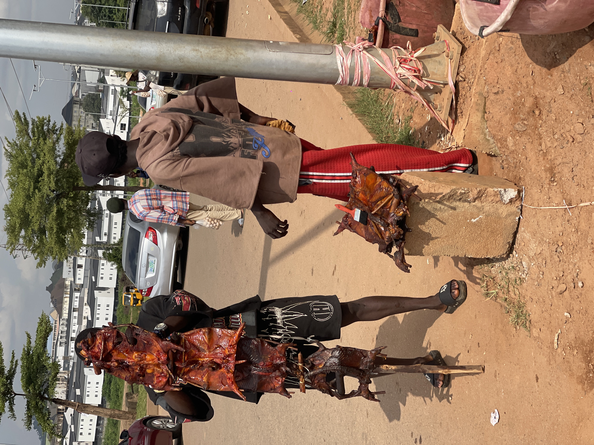 Two men are standing on a dirt road in Abuja, Nigeria. The man on the left is wearing a black t-shirt and black shorts, and he is holding a pole with several cooked animals on it. The man on the right is wearing a brown hoodie and red pants, and he is standing next to a concrete block with a cooked animal on it. In the background, there are several buildings and a silver car. The overall scene appears to be a street market or roadside stall selling cooked food.