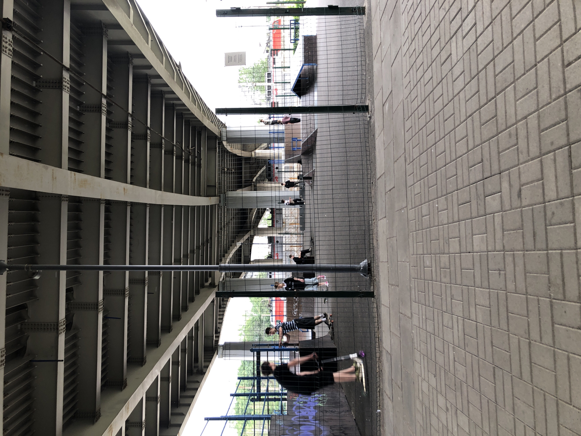The image shows an outdoor skate park area under a large bridge structure. Several young people are present. In the foreground, a person on a scooter rides from left to right. Further back, another individual stands on a skateboard. A group of people are gathered near a ramp structure, and one person is seated on a bench. A red train is visible in the background, suggesting an urban environment. The ground is paved with cobblestones, and a wire mesh fence separates the skate park from the foreground. The overall scene appears to be a public recreational space in Saint Petersburg, Russia.