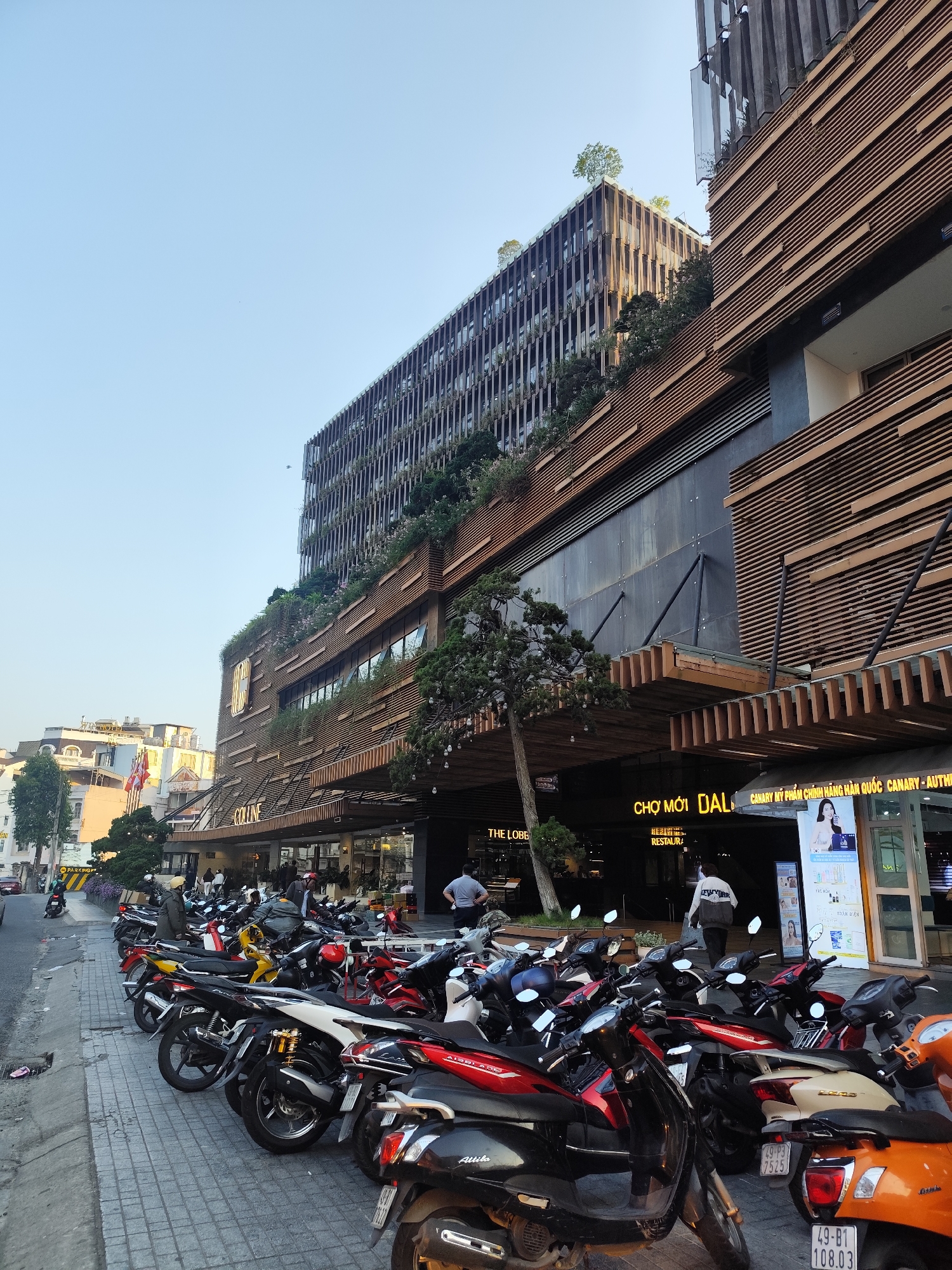 The image shows a street scene in Da Lat, Vietnam. In the foreground, many parked motorbikes occupy the sidewalk. Behind the motorbikes is the facade of a large building with multiple stories and a modern design, incorporating horizontal wooden slats and greenery. Signs above the entrance read "THE LOBBY" and "CHO MOI DAL", which identifies the building as the Dalat Center shopping mall. People are walking in and out of the entrance. The sky is pale blue, and other buildings are visible in the background.