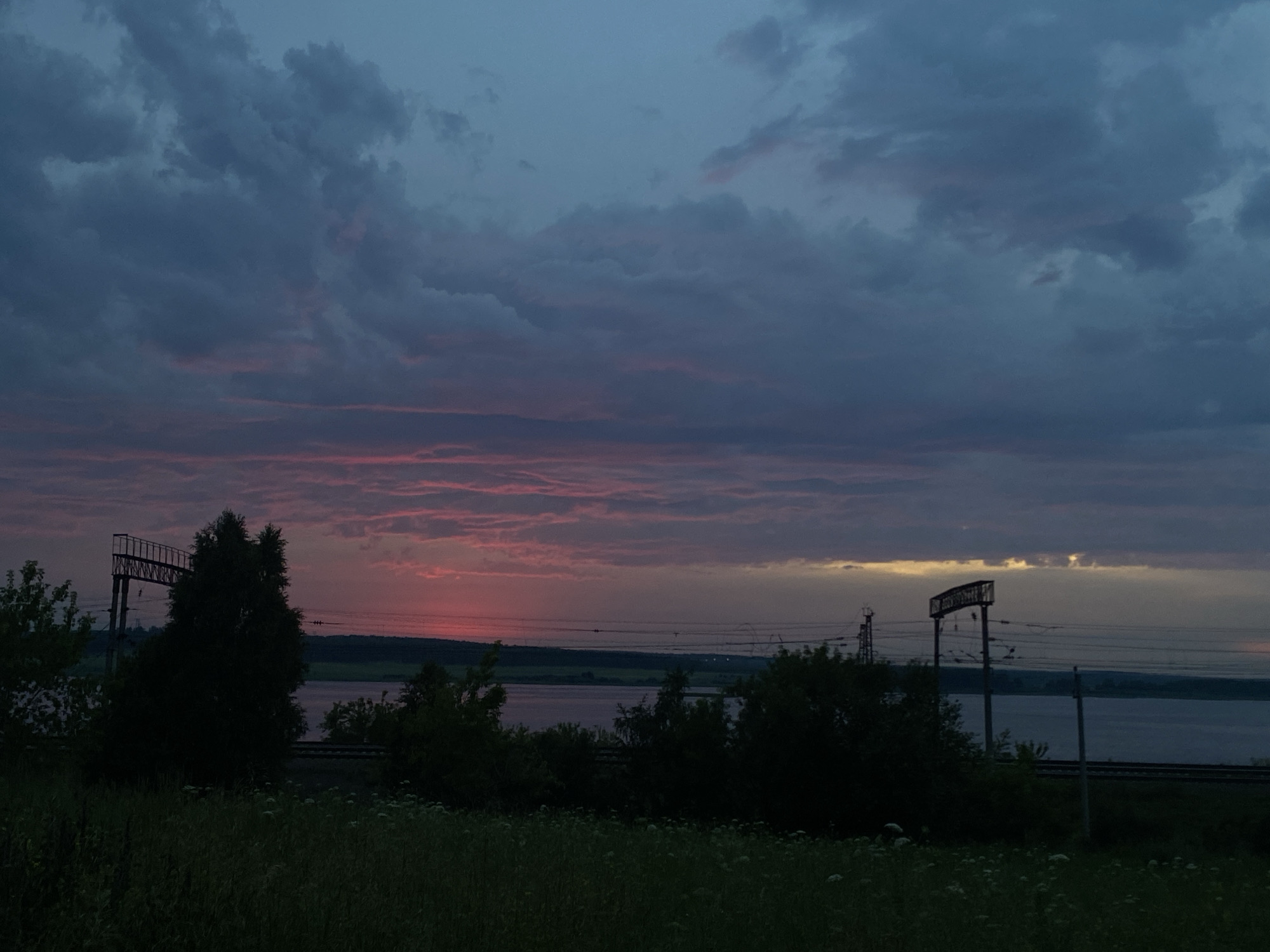 The image depicts a dusk scene in Smolino, Russia. The sky is a gradient of deep blue at the top, transitioning to hues of pink and orange near the horizon, indicative of a sunset. Clouds are present across the sky, with some illuminated by the setting sun. In the foreground, tall grass and wildflowers are visible. Beyond the grass, a body of water, likely a river or lake, stretches across the middle ground. Trees and vegetation line the banks of the water. Power lines and electrical poles are prominent features in the mid-ground, suggesting proximity to infrastructure. A large metal structure, possibly part of a bridge or industrial equipment, is visible on the left side of the frame. The overall atmosphere is serene and natural, with hints of human development.