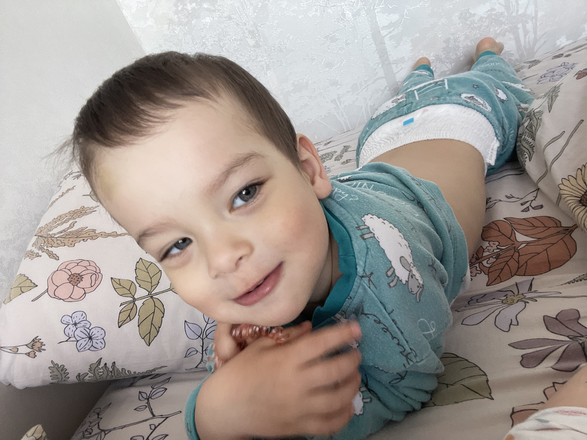 An indoor, eye-level shot features a young child, likely a toddler or preschooler, lying on their stomach on a bed or soft surface. The child, with short dark brown hair and striking blue-green eyes, looks directly at the camera with a gentle, engaging expression and a hint of a smirk. Their cheeks are flushed with a healthy color.
They are dressed in teal-colored, long-sleeved pajamas adorned with white sheep illustrations. A white pull-up diaper, possibly Huggies brand with a visible blue wetness indicator, is worn above the pajama pants. The child holds a coiled, multi-colored hair tie or bracelet in their hands, positioned near their chin.
The child rests on patterned bedding featuring various floral and leaf designs in soft hues of pink, blue, and brown. A matching pillow supports their head. In the background, a light-colored wall with subtly textured wallpaper, possibly depicting a frosted or natural tree-like pattern, is visible. The soft, ambient lighting suggests it is daytime indoors. The image conveys a sense of peaceful playfulness. No explicit visual cues in the image confirm the location as Izhevsk, Russia.