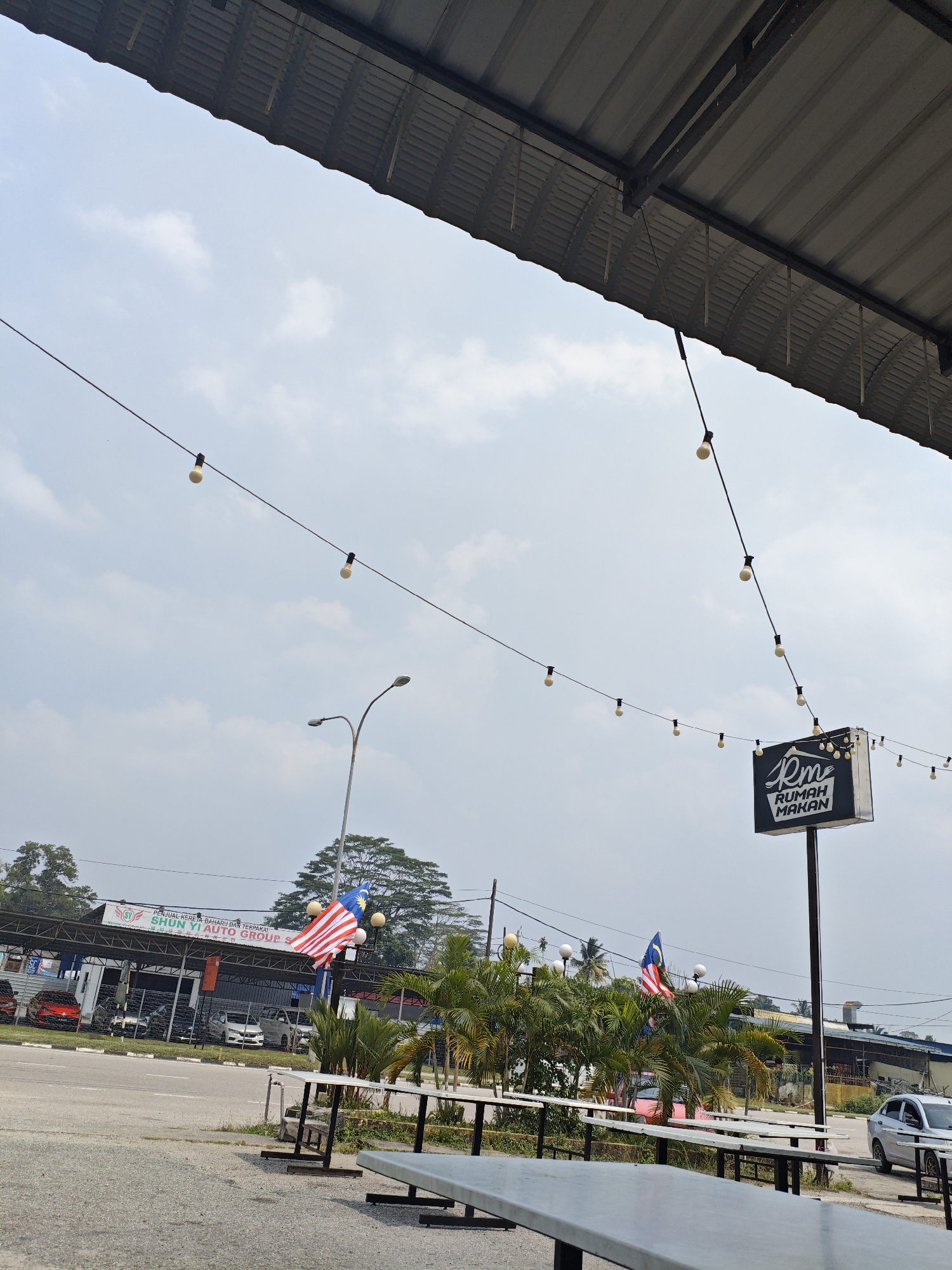 The image depicts an outdoor scene in Kota Tinggi, Malaysia, under a cloudy sky. In the foreground, there are several long, empty picnic tables made of grey material with metal legs. Behind the tables, a road is visible, with several cars parked on the side. Further back, a row of buildings with awnings is situated, one of which appears to be a car dealership with "SHUN YI AUTO GROUP" signage. A Malaysian flag is prominently displayed, attached to a pole in front of the buildings. String lights with visible bulbs hang across the scene, extending from a covered structure in the upper part of the image. A sign that reads "Rm RUMAH MAKAN" is also visible, indicating a place to eat. A street lamp stands to the left of the Malaysian flag. Lush green palm trees and other foliage are interspersed throughout the scene. The overall impression is a casual outdoor dining or rest area near a commercial establishment.