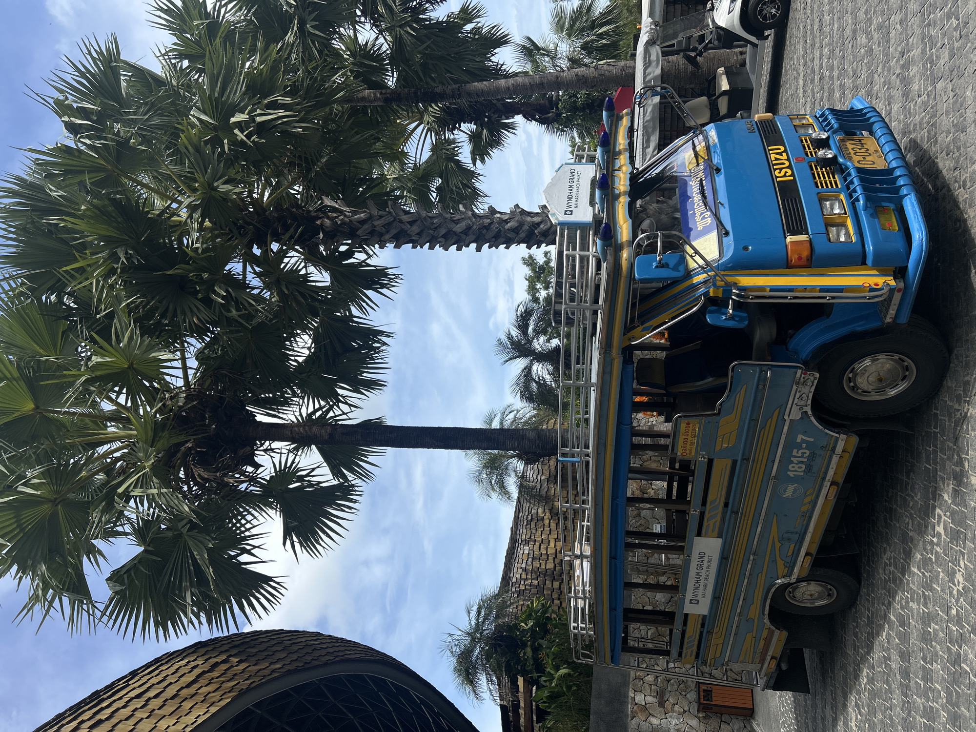 The image shows a blue Isuzu Songthaew bus, a type of converted pickup truck or van used as a shared taxi in Thailand, parked near palm trees. The bus has yellow accents and Thai script on the side. A Wyndham Grand Nai Harn Beach Phuket sign is attached to the side of the bus. A building with a brown, tiled roof is also visible. The location is likely in Rawai, Thailand. The sky is blue with scattered clouds. The ground is paved with gray stones.