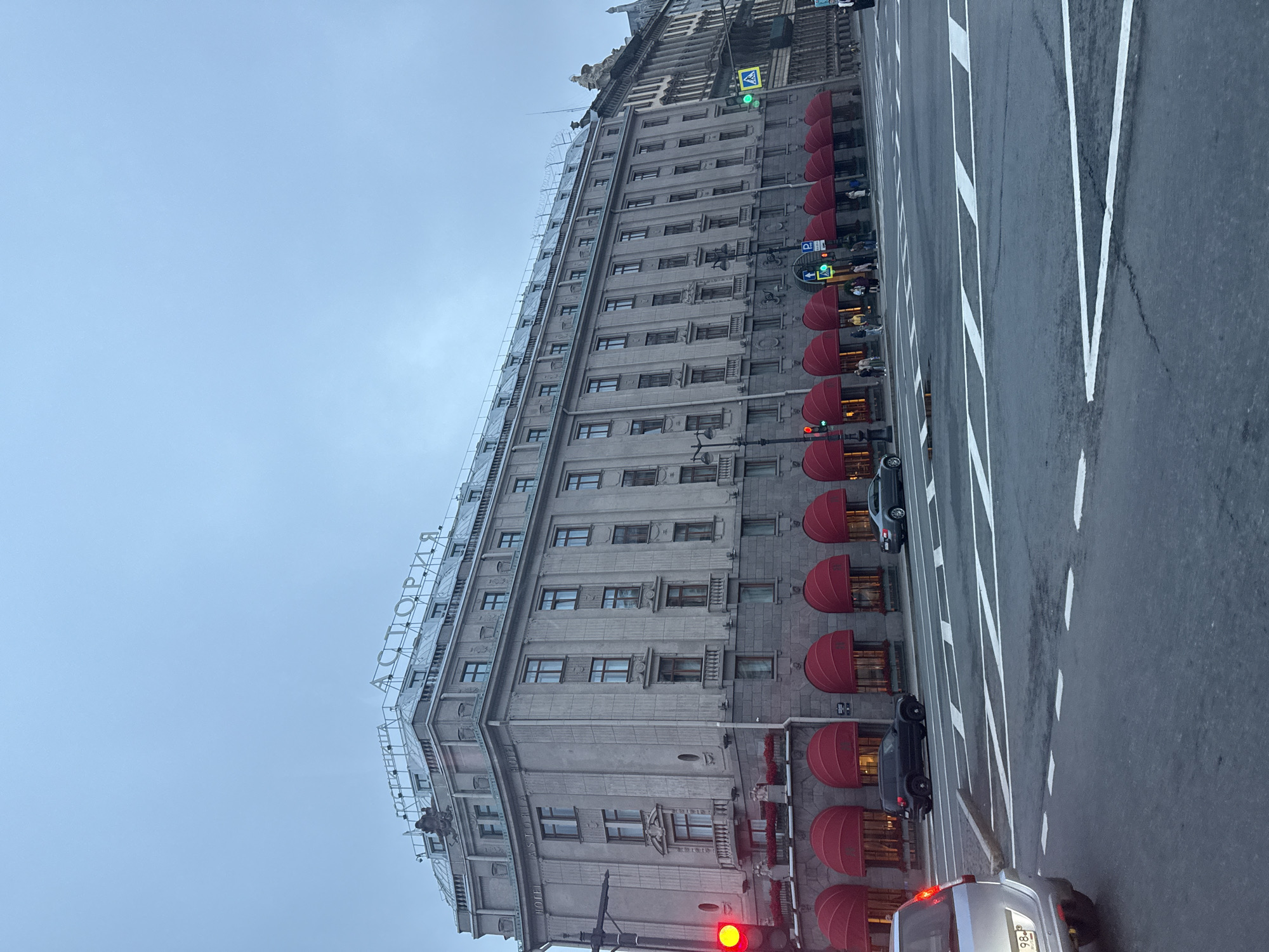The image captures a wide shot of the Astoria Hotel in Saint Petersburg, Russia, under an overcast sky. The grand, neoclassical building dominates the frame, its grey stone facade adorned with classical architectural details. Red awnings extend over the ground-floor windows, adding a splash of color against the muted tones of the building and sky. A prominent sign with Cyrillic letters, "АСТОРИЯ" (Astoria), is visible at the top of the building.
In the foreground, a street scene unfolds. Several cars are present: a silver SUV is parked on the left, and a dark SUV and a silver sedan are driving on the road. Pedestrians are visible on the sidewalk in front of the hotel. Traffic lights show red on the left, and a green light is visible further down the street. A pedestrian crossing with white lines marks the intersection. The road surface appears wet, suggesting recent rain. The overall atmosphere is one of an urban environment on a subdued, possibly cool, day.