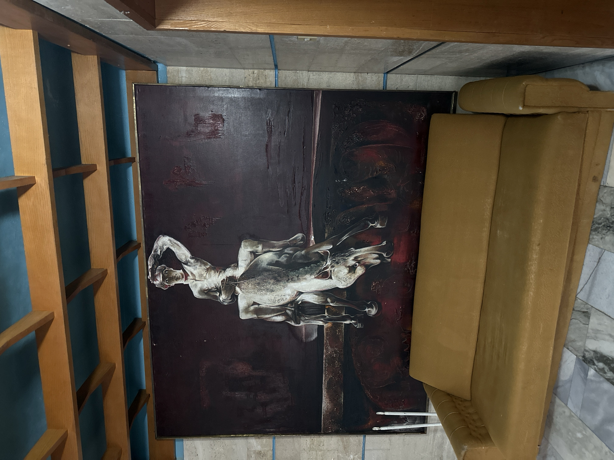 The image shows an interior space. On the left, there's a wooden bookshelf structure with blue backing. A large painting dominates the center, depicting what appears to be a dramatic scene with a figure on horseback in a dark, abstract style. To the right of the painting, there is a large tan-colored upholstered structure, possibly a couch or sectional, partially visible. The surrounding walls appear to be a neutral color, with some blue cable management visible. The floor is made of light marble. The location is unknown.