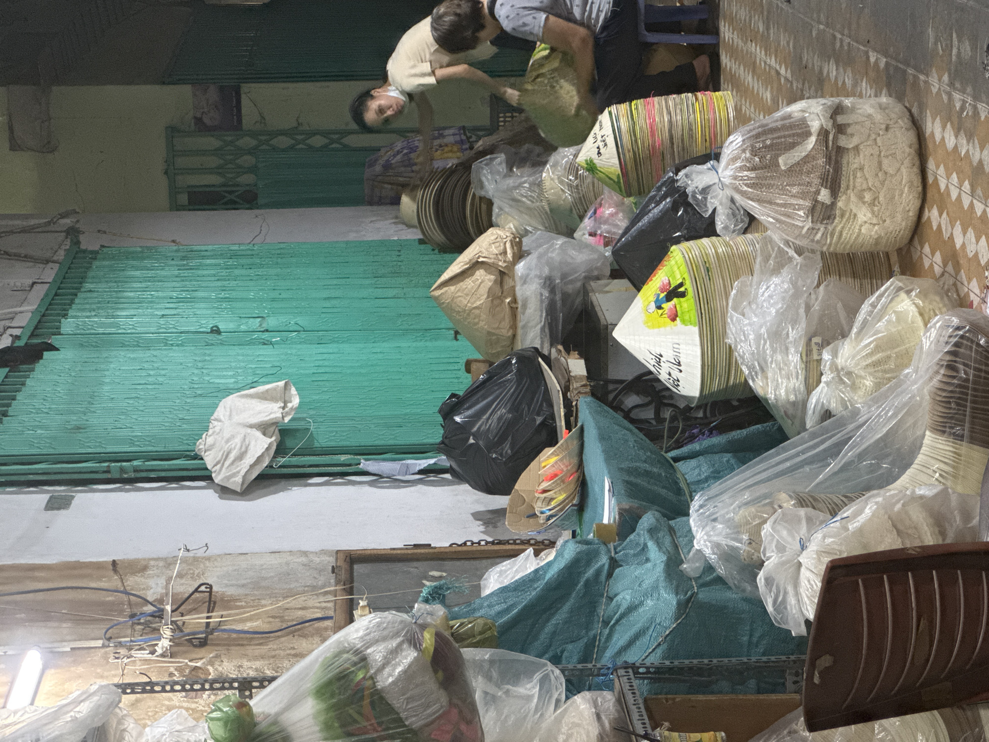 This image depicts an outdoor market stall in Nha Trang, Vietnam. The stall is filled with a variety of conical hats, some individually wrapped in plastic, others stacked high in colorful arrangements. In the background, two women are visible. One woman, wearing a light-colored shirt and a face mask, appears to be arranging or sorting materials. Another woman, seated on a blue stool, is holding a large, light-green bag. The stall is characterized by its cluttered arrangement of goods, plastic sheeting draped over items, and a general atmosphere of a busy marketplace. The green corrugated metal gate behind the stall suggests a permanent structure, possibly a storefront or a covered market area. The ground is tiled with a diamond pattern. Electrical wires and a metal shelving unit are also visible, contributing to the utilitarian and somewhat makeshift appearance of the stall. A single white garment hangs on the wall near the gate. The lighting suggests it is either early morning or late evening.