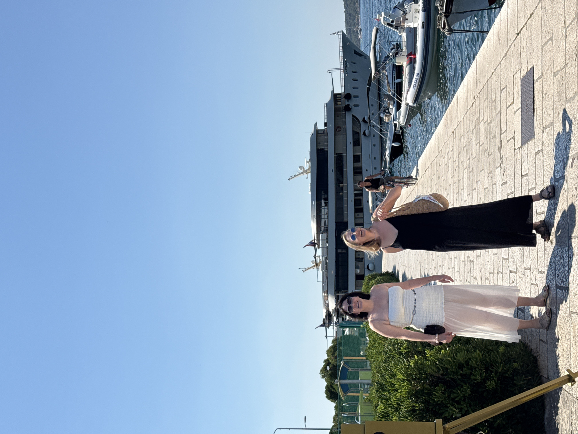 Two women are standing on a stone promenade in the city of Šibenik, Croatia, with a large yacht and several smaller boats docked in the harbor behind them. The woman on the left is wearing a white strapless dress with a sheer overlay and sunglasses. The woman on the right is wearing a black slip dress and sunglasses, carrying a straw bag. A person is cycling on the promenade in the background. The sky is clear and blue, and the water is calm. Green bushes line the promenade to the left.