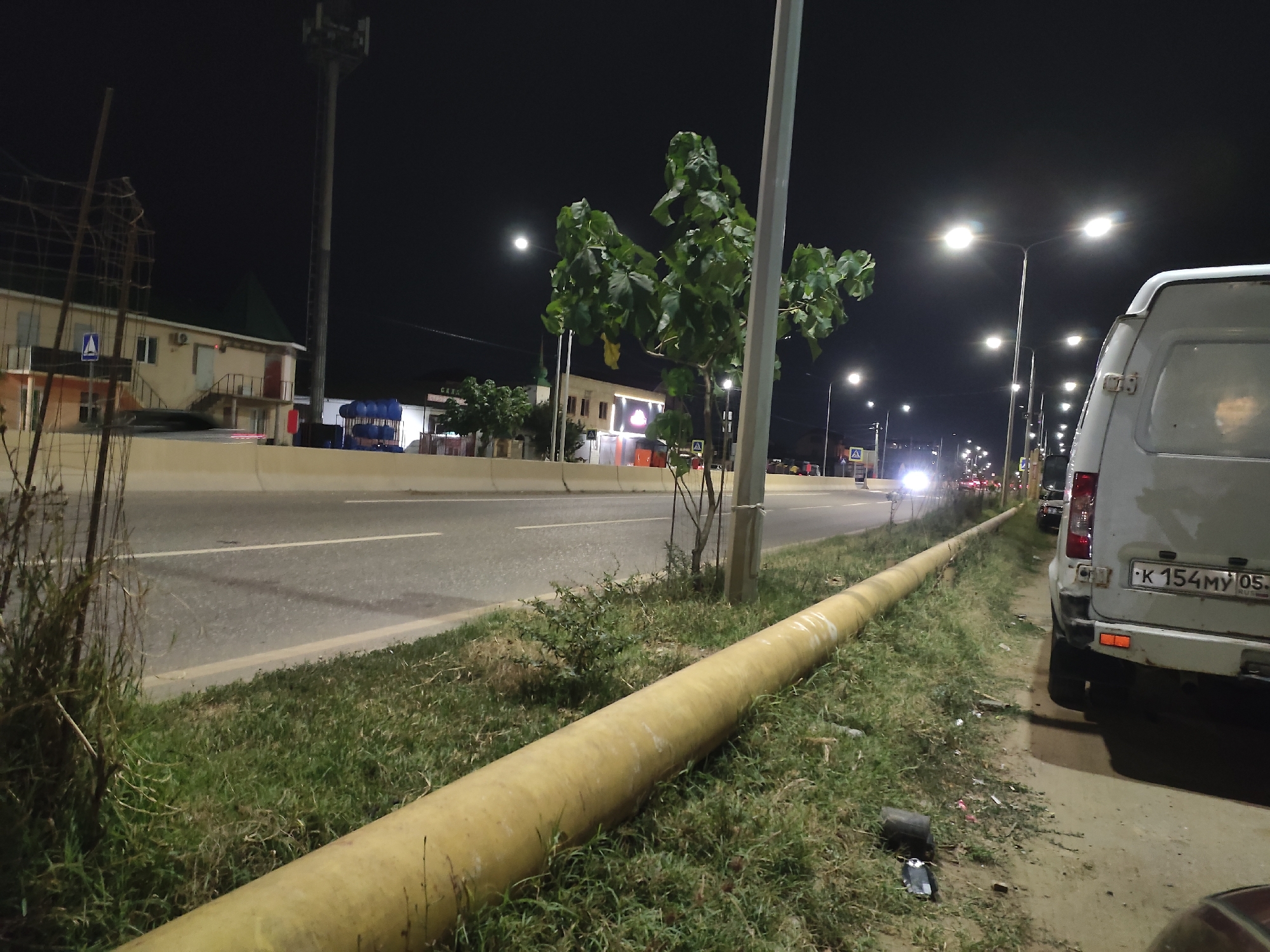 At night, a wide, paved road with a white stripe runs horizontally across the image. On the left side of the road, a grassy median is present, and a large, yellow pipe runs along the edge of the median. Beyond the median, buildings and a cell tower are visible. On the right side of the road, a white van is parked, with its rear end facing the viewer. The van has a license plate that reads "K 154MY 05". The scene is illuminated by streetlights and the headlights of passing vehicles. The environment appears to be urban, with the city of Leninkent, Russia, being the implied location. There are no people visible in the image.