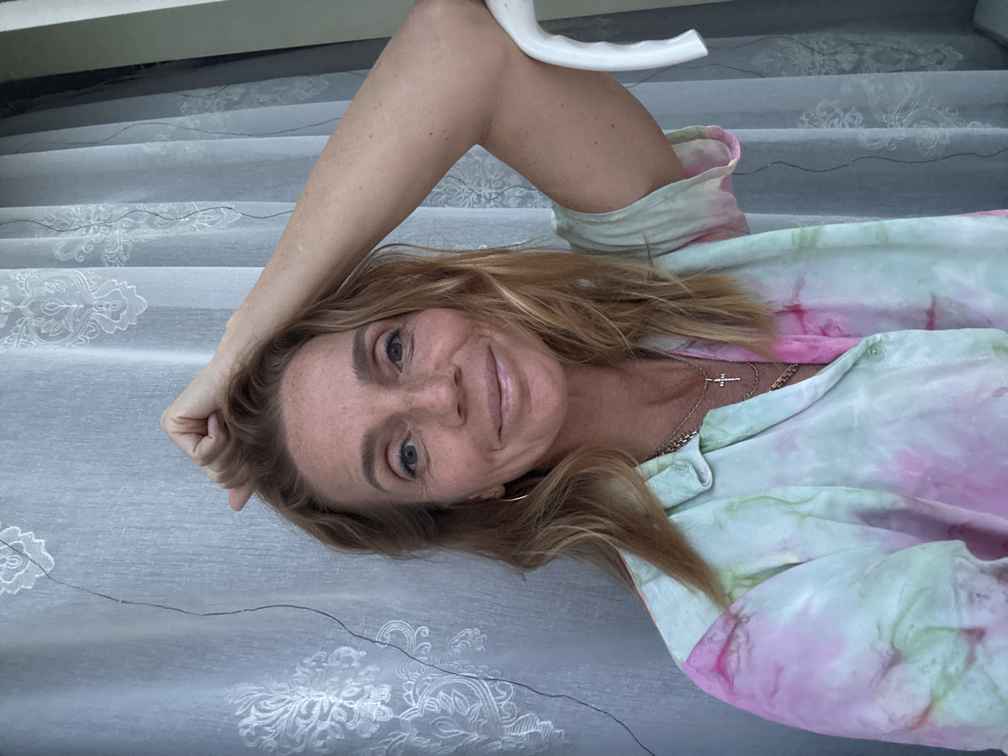 A woman is pictured from the chest up, in a self-portrait. She has blonde hair, freckles and blue eyes. She is wearing a tie-dye shirt in shades of pink and green. Her right arm is raised and bent, with her hand on her head. She is leaning on a white object, possibly a chair. Behind her, a sheer, light blue curtain with a white embroidered pattern is visible. The scene is indoors, possibly a home, with no specific city or country identifiable.