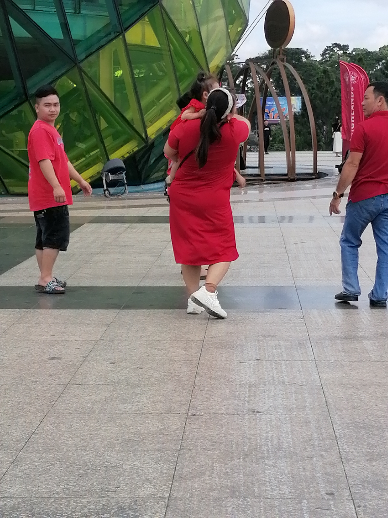 The image depicts an outdoor scene in the city of Da Lat, Vietnam, likely at a public space with a modern architectural element. In the foreground, a man wearing a red t-shirt and dark shorts walks on a paved ground made of square tiles. To his right, a woman in a vibrant red dress is carrying a young child. The woman is facing away from the camera, and the child is positioned on her back, also facing away. Further to the right, another man, also in red, is seen from the side, wearing blue jeans and a watch. Behind the group, a large, multifaceted green structure with geometric shapes dominates the background, possibly a building or an art installation. The sky is visible above, with a hint of trees and a red flag with white lettering on the right side. A metal sculpture with curved elements stands in the middle ground.