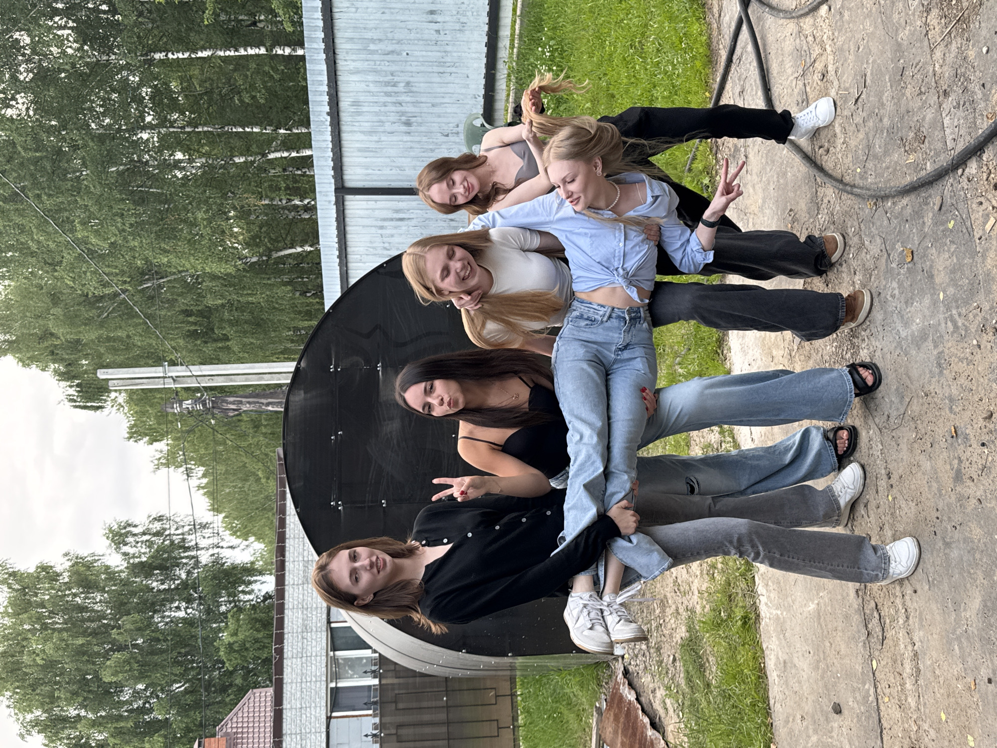 The image shows a group of five young women posing together outdoors. They are arranged vertically in the frame, with the person at the top closest to the camera and the person at the bottom furthest away. The setting appears to be a backyard or outdoor recreational area, with grass, trees, and a concrete surface visible. There's a large, black, circular object in the background that resembles a satellite dish. In the far background, a wooden fence and some buildings are visible.
The women are dressed casually in jeans, t-shirts, and a light blue collared shirt. One woman is wearing a pearl necklace. Some of the women are smiling and interacting with each other, suggesting a friendly and relaxed atmosphere. One woman is making a peace sign with her fingers.
The lighting suggests it might be daytime, possibly late afternoon given the soft shadows. The weather appears to be overcast or cloudy. The location context indicates Perm, Russia, but there are no specific text-based clues within the image to confirm this.