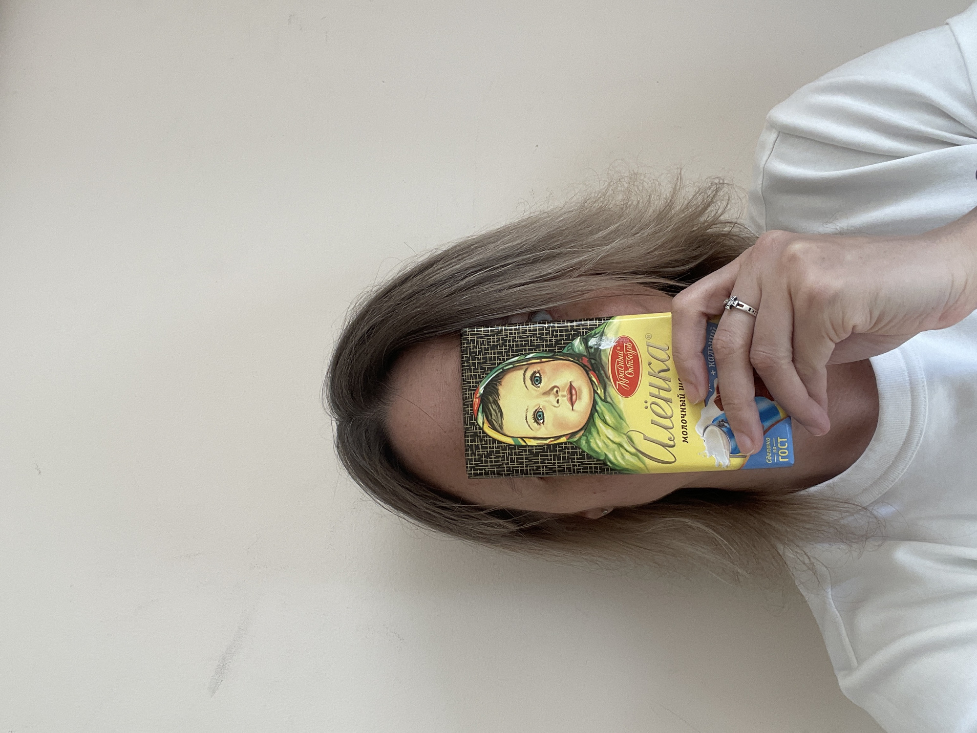 A woman with blonde hair is holding a yellow chocolate bar in front of her face. The chocolate bar has the image of a young girl wearing a yellow headscarf. The woman is wearing a white t-shirt and a ring on her finger. The background is a light beige wall. The woman is likely in Perm, Russia, because the chocolate bar is a Russian brand.