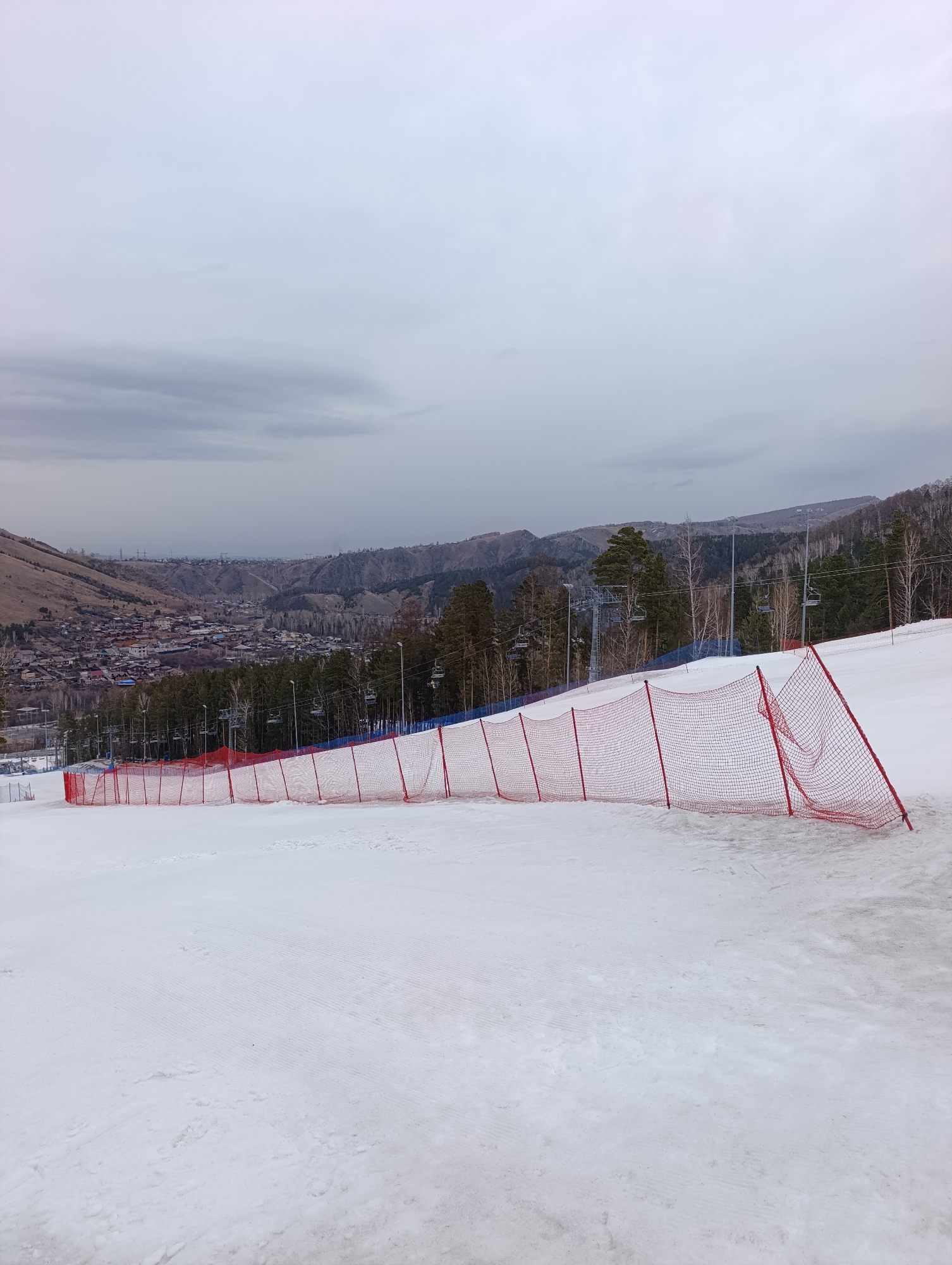 The image depicts a snowy ski slope with a red safety net running along its side. In the background, a town nestled amongst hills is visible under a cloudy sky. Ski lift infrastructure is also present. Based on these details and the prompt, it's possible this scene is near Krasnoyarsk, Russia. The snow appears groomed, suggesting active use of the slope. No people are immediately visible in the image.