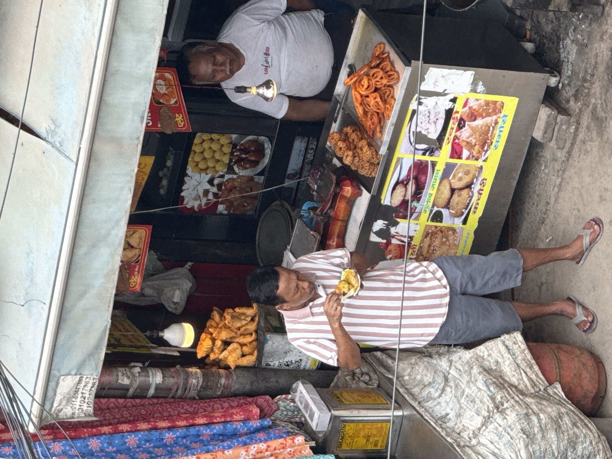 The image captures a street food stall in Delhi, India, likely in a busy marketplace given the close proximity of buildings and the presence of various goods. A man in a striped polo shirt and grey shorts is standing and eating from a plate held in his hand, using a spoon. He appears to be a customer or an employee. Behind him, a second man, wearing a white t-shirt with "Unlimited" printed on it, stands behind the counter, seemingly serving or observing.
The stall displays a variety of fried snacks and sweets. In the foreground, a tray holds numerous circular, orange-colored sweets, possibly jalebis, and another tray contains intricately shaped fried items. A large display board with images of different food items, including samosas, is affixed to the front of the stall. There are also piles of fried pastries visible on a higher shelf.
The environment is typical of an Indian street food setting, with an awning providing shade, a hanging lightbulb illuminating the food, and various containers and goods stacked around. Fabric drapes, possibly from a nearby shop, are visible on the left. The ground appears to be concrete or pavement. The overall impression is a snapshot of daily life and commerce in a bustling urban environment.