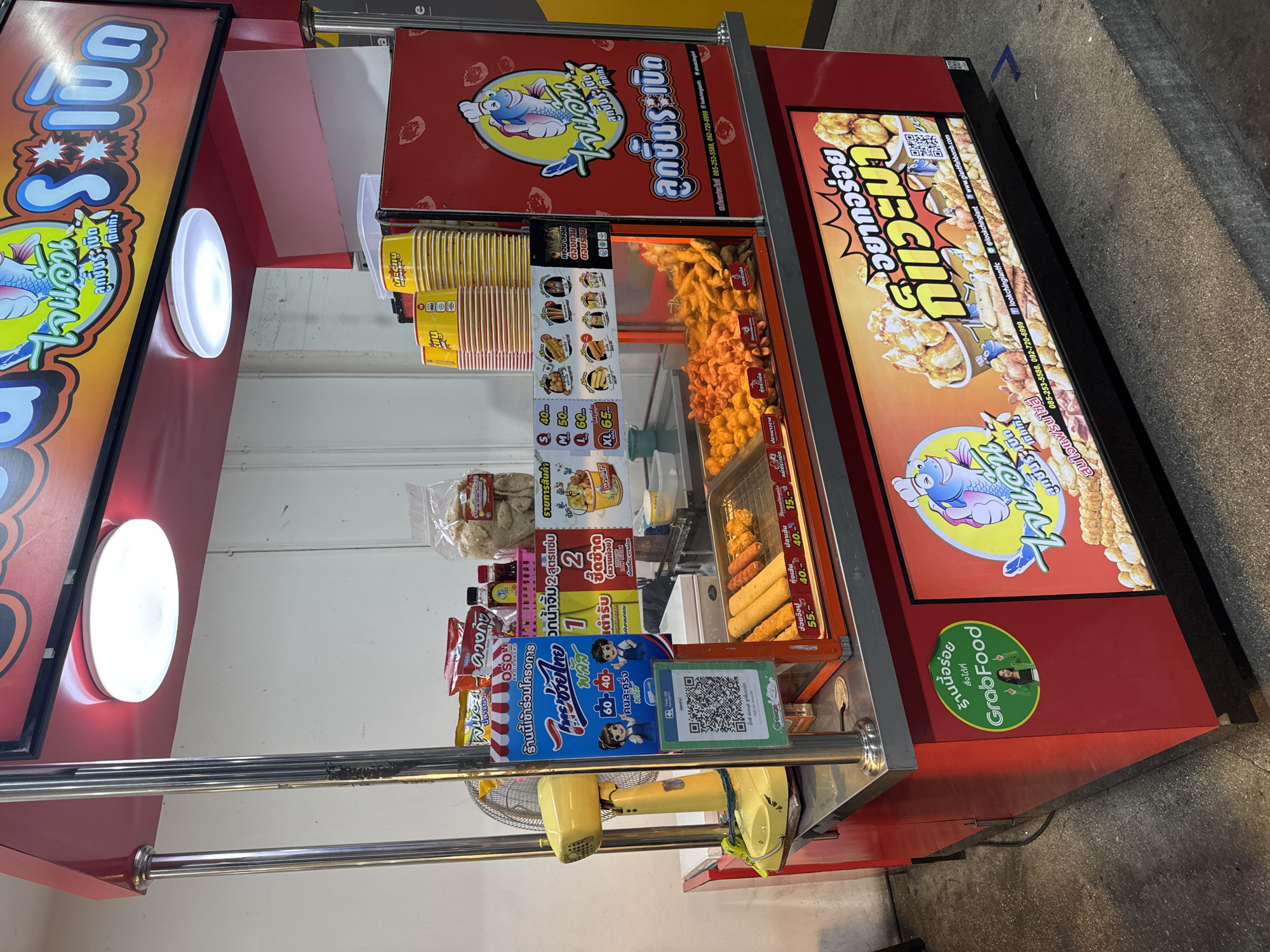 This is a street food stall in Bangkok, Thailand, specializing in fried fish balls. The stall is bright red and yellow, with a large sign in Thai featuring cartoon fish characters and the name of the stall, "Look Ching Tantee" (ลูกชิ้นเต้นท์). The food is displayed in a glass case, with various fried items, including fish balls, chicken nuggets, and sausages, available for purchase. There are also bags of snacks and drinks on display. A QR code is visible on the stall, likely for payment or ordering. The ground in front of the stall is concrete, and there is a yellow fan on the counter. A GrabFood sticker is also present, indicating delivery services.