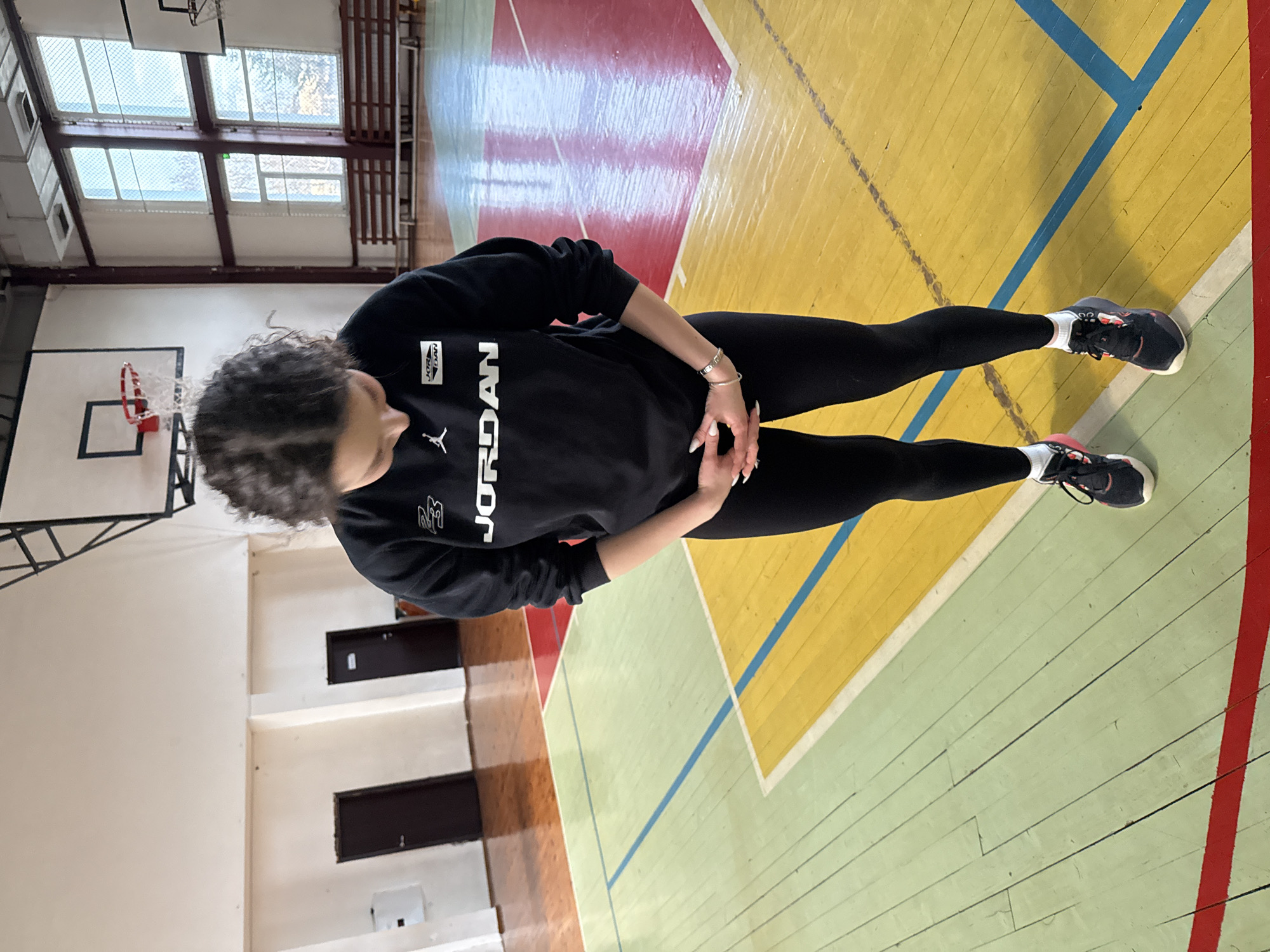 A young female with dark, curly hair stands within a gymnasium, her head bowed and hands clasped in front of her waist. She is dressed in a black Jordan brand sweatshirt with white lettering and logos, black leggings, and black and pink athletic shoes with white socks. Her fingernails are painted white, and a silver-toned bracelet is on her left wrist.
The gymnasium floor is constructed of wood, painted in distinct sections of red, yellow, and light green, marked with blue and red court lines. The floor shows signs of wear but is reflective. In the background, light-colored walls are visible. On the left wall, two dark brown doors are present, one featuring a white pictogram. On the right, large multi-paned windows provide natural light, revealing bare trees and horizontal wooden benches outside. Two basketball backboards with red hoops and nets are visible; one is prominent in the mid-ground to the right, and another is partially seen on the far left. Metal support structures are visible near the ceiling.
This scene takes place within a sports hall consistent with a public school or community gymnasium in the city of Sofia, Bulgaria. The precise location within Sofia is not identifiable from the provided visual information.