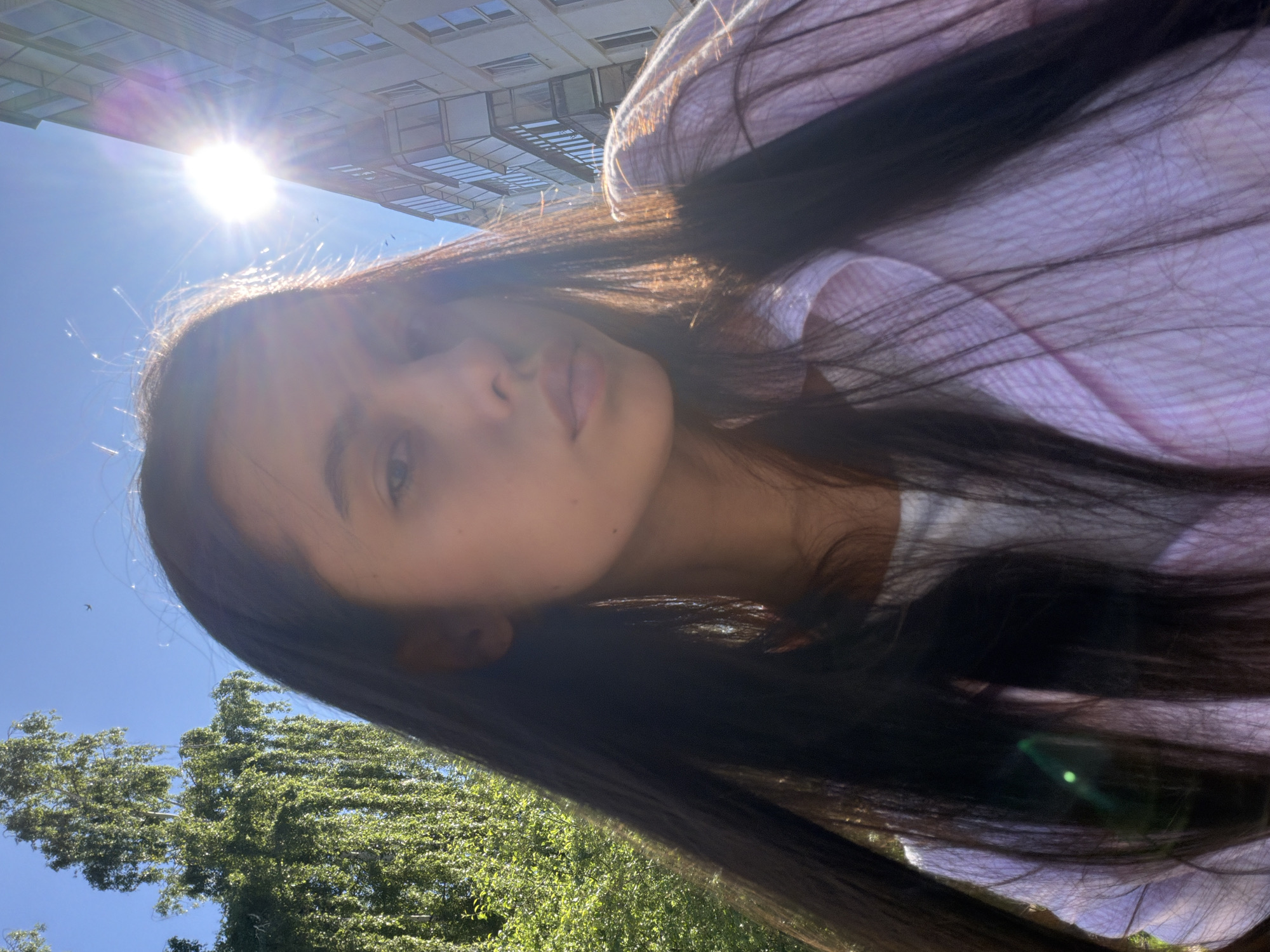 A young woman with long, dark hair is captured in a selfie taken outdoors. She is wearing a pink striped collared shirt over a white undershirt. The sun is bright, creating a lens flare and casting a warm glow on her face and hair. In the background, a large tree with lush green leaves is visible on the left, and a multi-story building with balconies is on the right. The sky is clear and blue. The image appears to be taken during the daytime. The presence of trees and a residential building suggests an urban or suburban environment. Chaikovsky, Russia is a city known for its industrial facilities and natural beauty, with many green spaces and proximity to the Kama River. This scene is consistent with an outdoor setting in such a city.