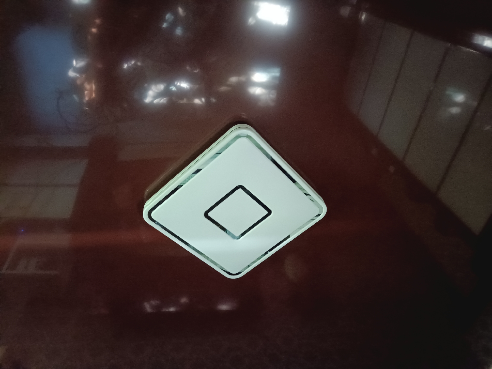 A diamond-shaped object with a white outer rim and a light blue inner rim is centered on a dark, reflective surface. Inside the diamond shape, a smaller black diamond outline encloses a white space. The reflective surface shows blurred light patterns and hints of objects in the background, including what appears to be a bicycle and some vertical rectangular shapes. The overall impression is that of a modern, sleek object placed on a polished, possibly furniture, surface. The context of Donetsk, Ukraine, is not evident from the visual information alone.