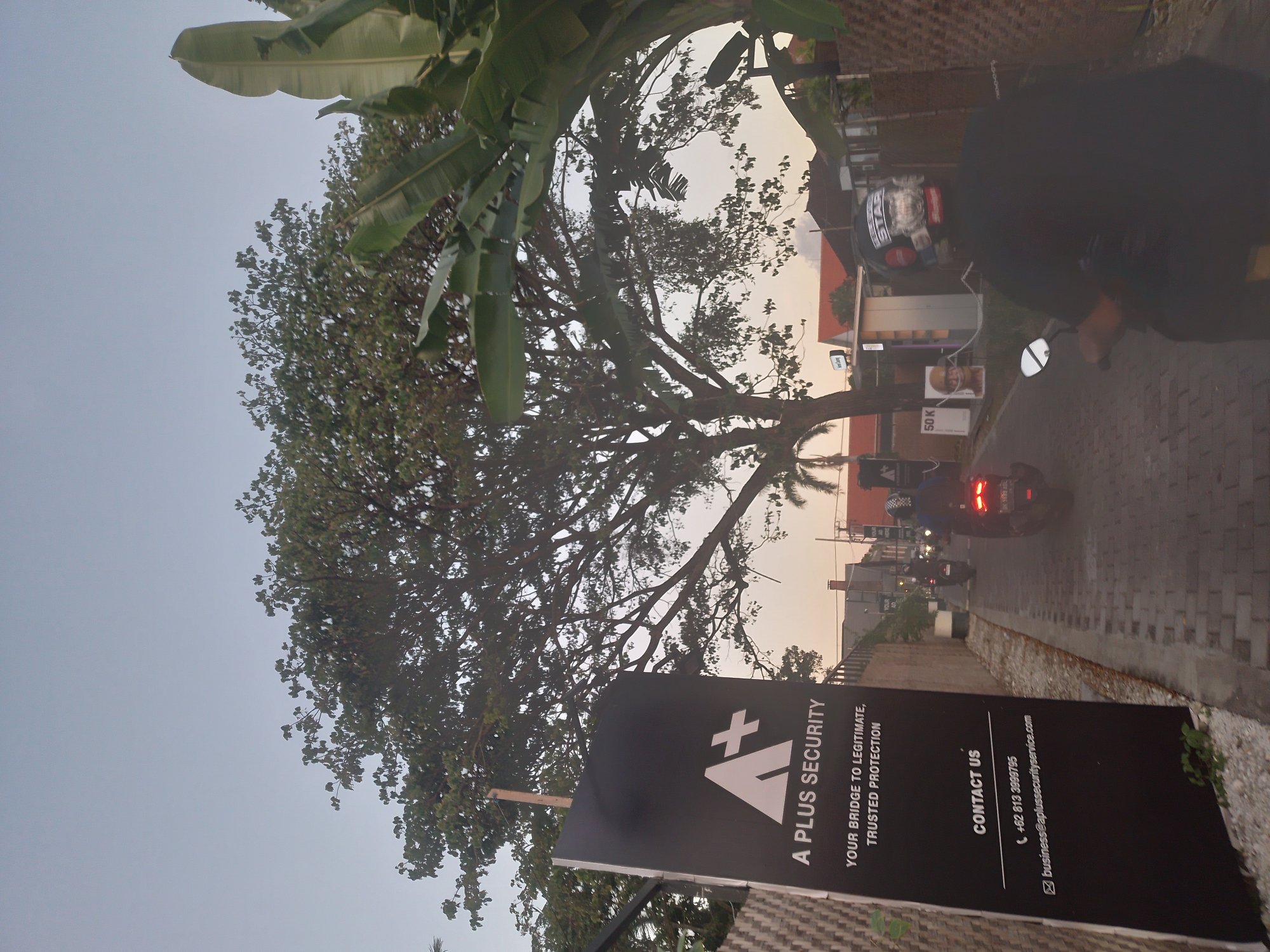 The image, taken in North Kuta, Indonesia, shows a street scene at dusk. A large tree and banana leaves dominate the left side of the frame. A black sign for "A Plus Security" is positioned in the foreground, displaying contact information. The street is paved with brick and is lined with buildings and greenery. Several motorbikes are visible on the street, with at least one actively driving away from the camera. A person on a motorbike is positioned on the right edge of the frame.