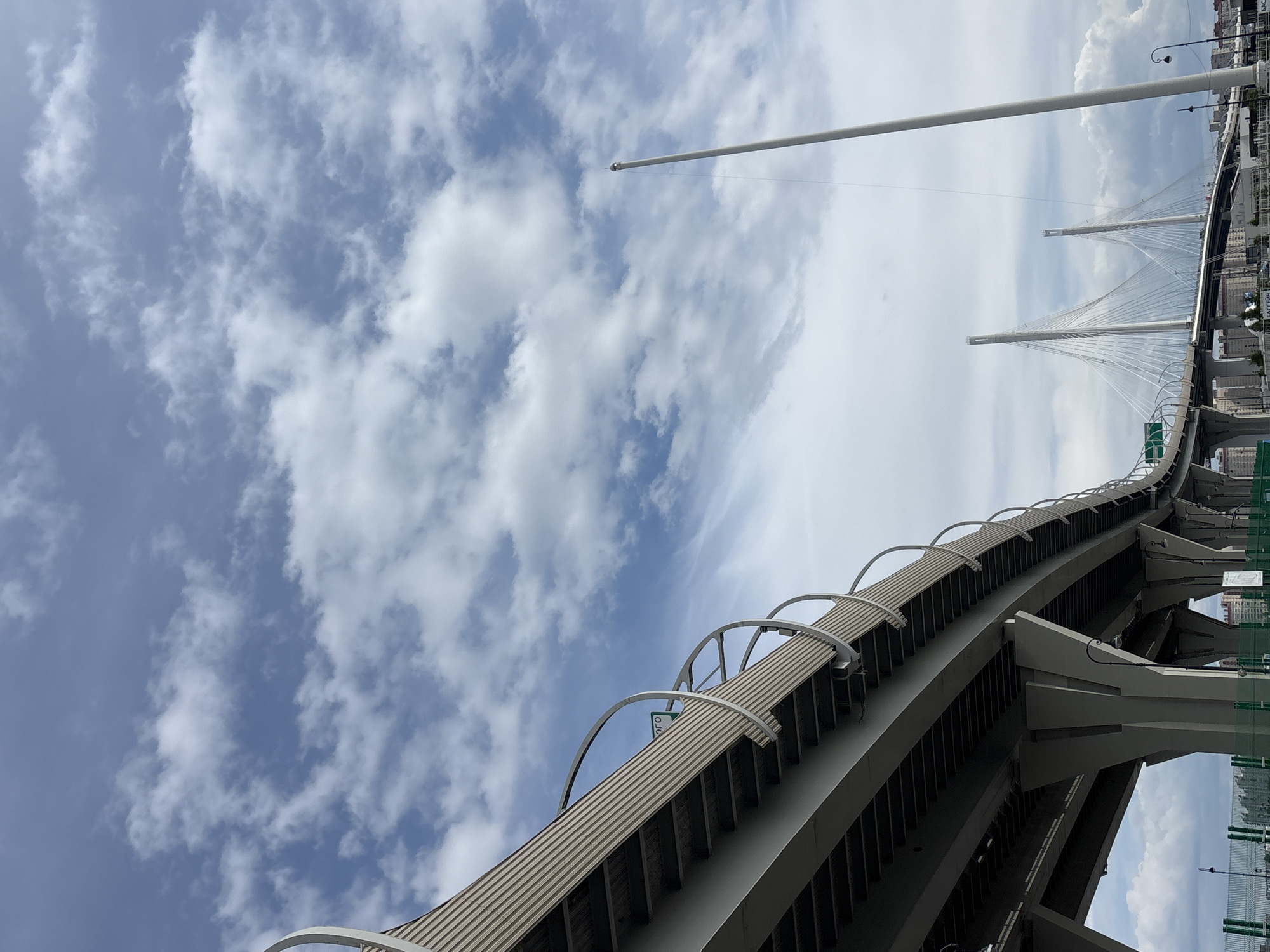 This image captures a vibrant daytime scene dominated by architectural infrastructure against a clear sky. The sky, occupying the upper left portion, is a bright blue adorned with scattered white cumulus clouds, indicating fair weather.
In the foreground, an elevated highway or ramp curves diagonally across the lower right of the frame, constructed from gray concrete and featuring a ribbed, metallic-looking surface on its upper deck. A series of distinctive white, arched guardrails line the outer edge of this structure. Its underside reveals complex concrete support beams.
Extending from the upper center towards the upper right is a tall, slender, light-colored pole, possibly a flagpole or part of the bridge's lighting system, terminating in a small, rounded cap.
In the background, positioned towards the right side of the image, stands a majestic cable-stayed bridge. It is characterized by two tall, white, modern pylons from which numerous white cables fan out gracefully to support the bridge deck below. Beyond the bridge, faint outlines of urban buildings are visible.
No people or discernible activity are present in the image, which focuses on the structural grandeur. The bright lighting suggests it is daytime, likely mid-morning or early afternoon.
Visible text includes a partial green sign on the elevated highway structure showing white Cyrillic characters, possibly "ого". In the far background, on banners near the cable-stayed bridge, Cyrillic text like "БАССТРИМ" is also visible. Given the context, this scene likely depicts a modern bridge infrastructure in Saint Petersburg, Russia.