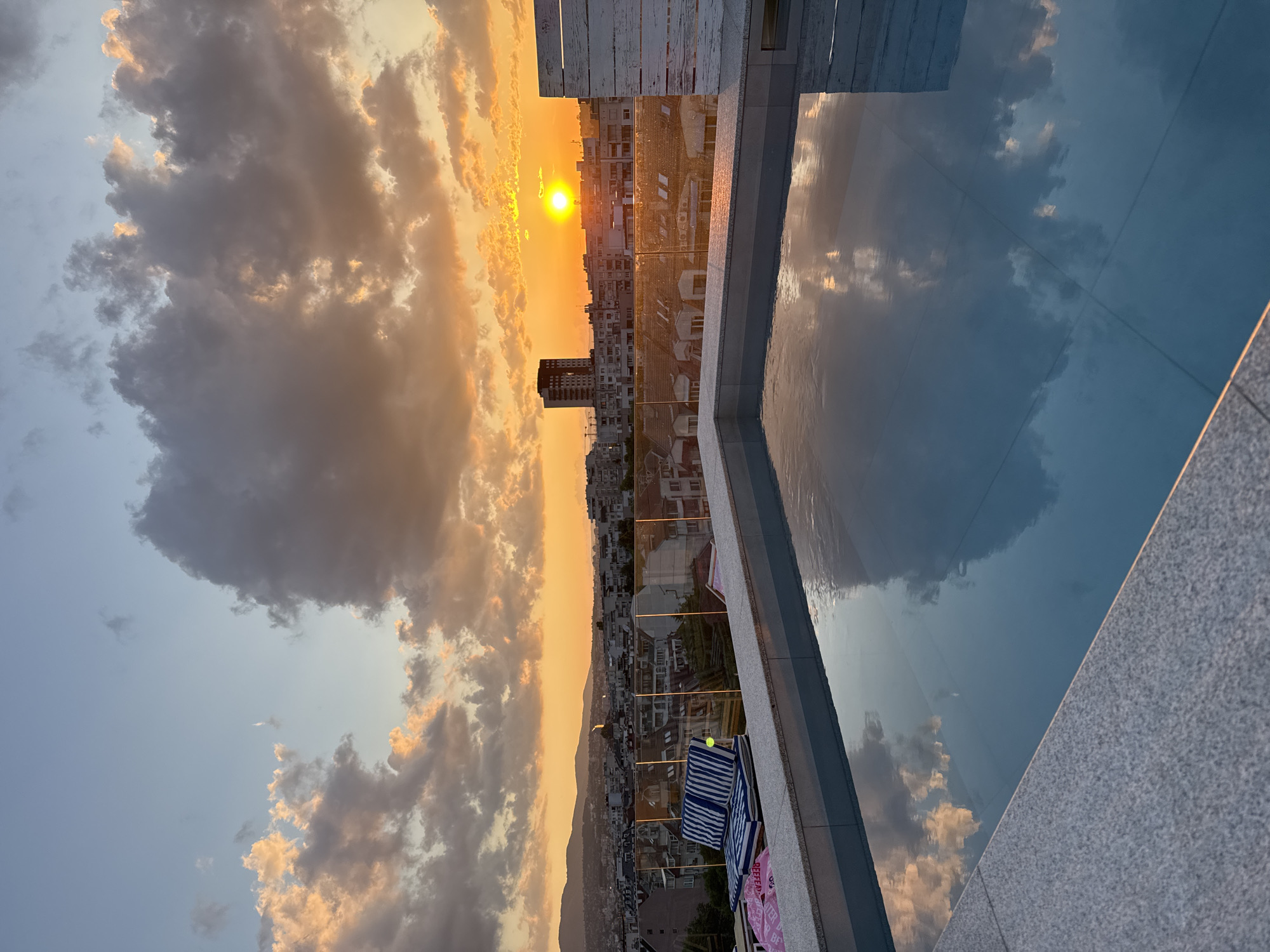 The image presents a rooftop perspective over the city of Sofia, Bulgaria, during a sunset. The lower half of the frame features a rectangular pool with tranquil, blue water reflecting the sky's colors and cloud formations. The pool is bordered by a light grey, speckled granite coping. Beyond the pool, a transparent glass barrier separates the water from an outdoor lounge area. A sun lounger with blue and white striped cushions is visible to the left, with a pink fabric item partially visible beneath it. To the far right, a white, vertically slatted wooden panel is present.
The midground and background depict a sprawling urban landscape of Sofia, characterized by numerous residential and commercial buildings of varying heights and architectural styles, extending towards a mountain range in the distant background, identifiable as the Vitosha mountains. A prominent, darker high-rise building stands out against the cityscape towards the center-right. The sky is dominated by a vivid sunset, with the sun appearing as a bright, circular yellow-orange disc positioned on the right horizon. Cumulus clouds are heavily present, displaying warm hues of orange, yellow, and reddish-brown where illuminated by the setting sun, contrasting with darker grey-blue areas. The upper portion of the sky is a lighter blue. No individuals are present in the scene.