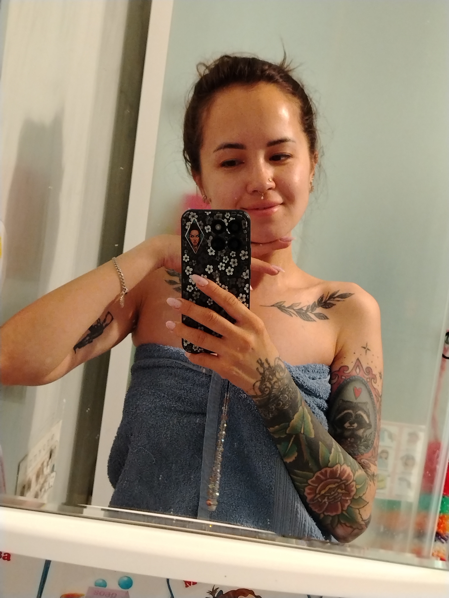 A medium shot depicts a woman indoors, seemingly in a bathroom, as she takes a selfie in front of a mirror. She has brown hair pulled back in a bun, light skin, and several tattoos. She wears a blue towel wrapped around her torso. The woman's right arm, heavily tattooed with floral and other designs, is visible. Her left arm has a smaller tattoo and a bracelet. She holds a cell phone with a patterned case, seemingly a selfie, in her right hand. There are various toiletries on a counter in the foreground, and a light blue wall is visible in the background. There is no indication of Noviy, Russia.