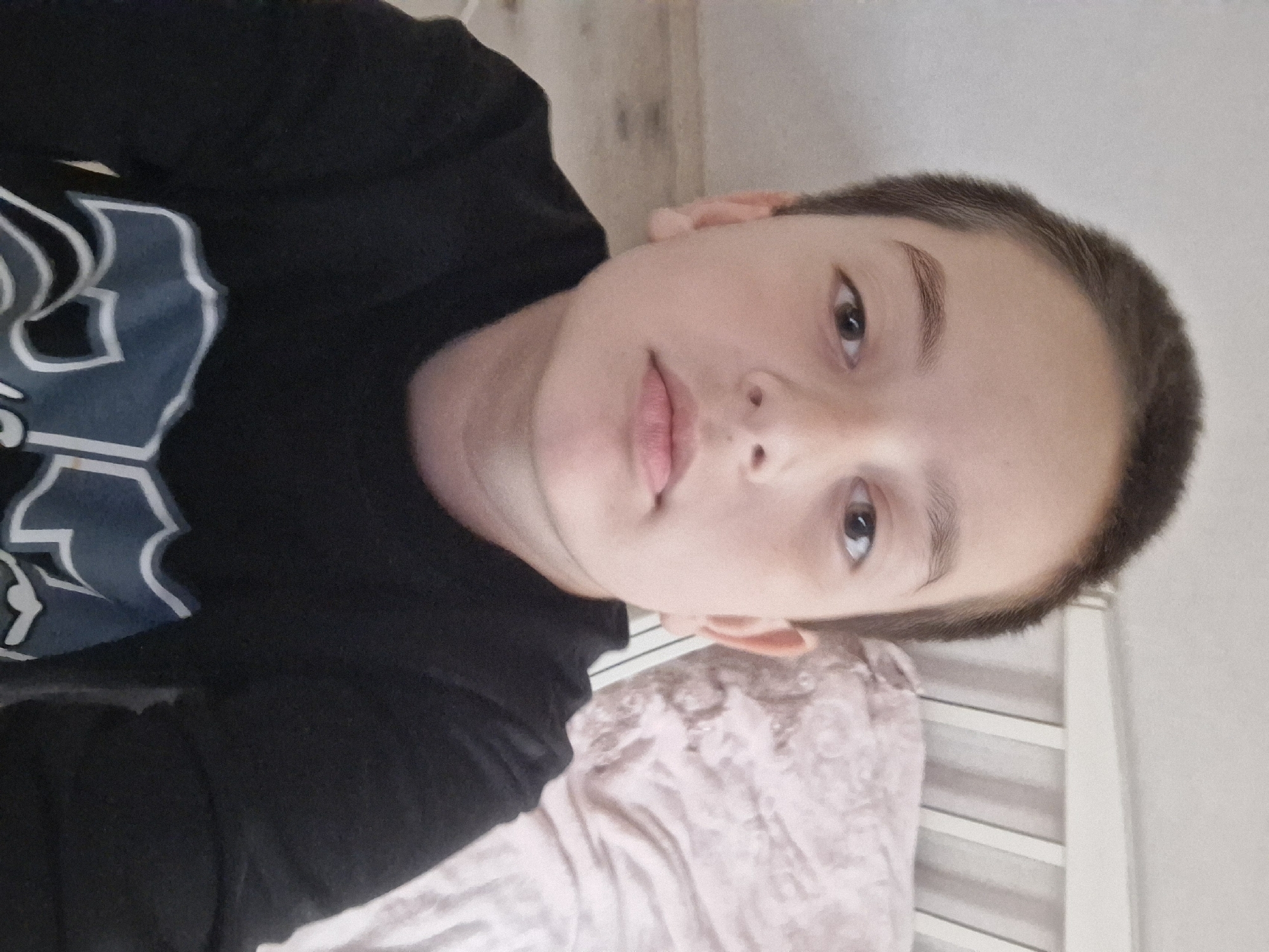 A young person with short brown hair, light skin, and brown eyes is positioned in the center of the frame. They are wearing a black long-sleeved shirt with white and blue lettering on the chest. The person appears to be lying on a bed with a pink textured blanket. A white headboard is visible in the background. The background is a light beige wall. The image is rotated to the side. It is not possible to determine if this image was taken in Shali, Russia or elsewhere based on the visible information.